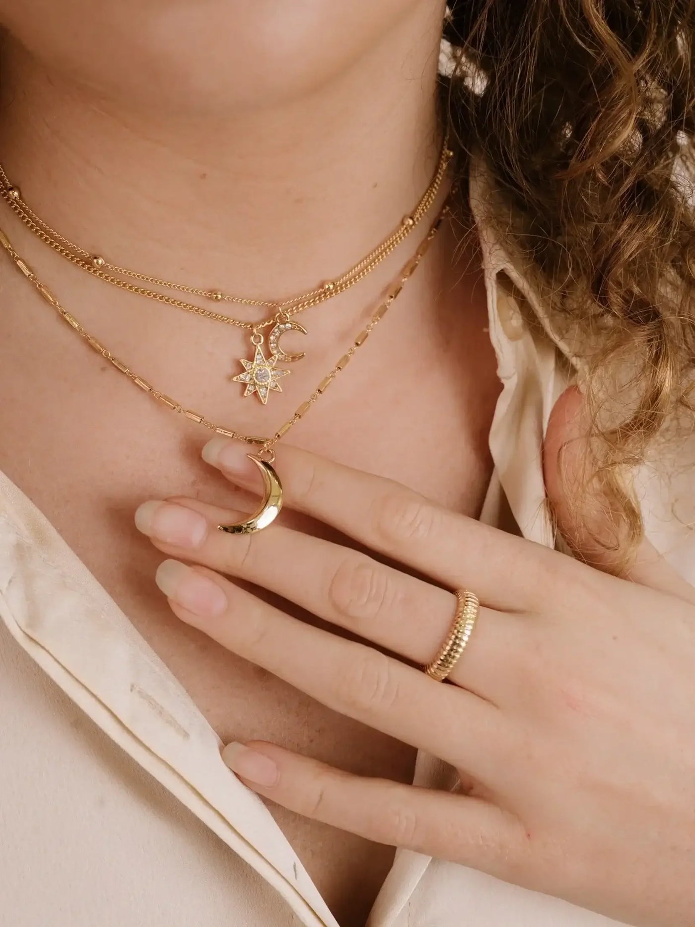 Gold jewelry adorns a neck, featuring layered necklaces with a crescent moon and star pendant. A hand with manicured nails gently touches the jewelry, set against a light-colored shirt.