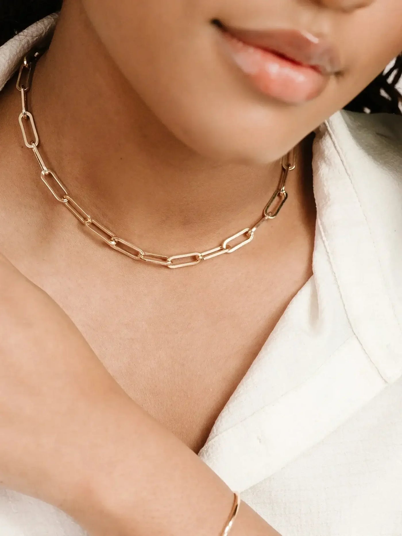 A gold paperclip-style necklace rests around a person's neck. The individual lightly touches their collarbone while wearing a white, textured shirt, creating an elegant appearance in a neutral setting.