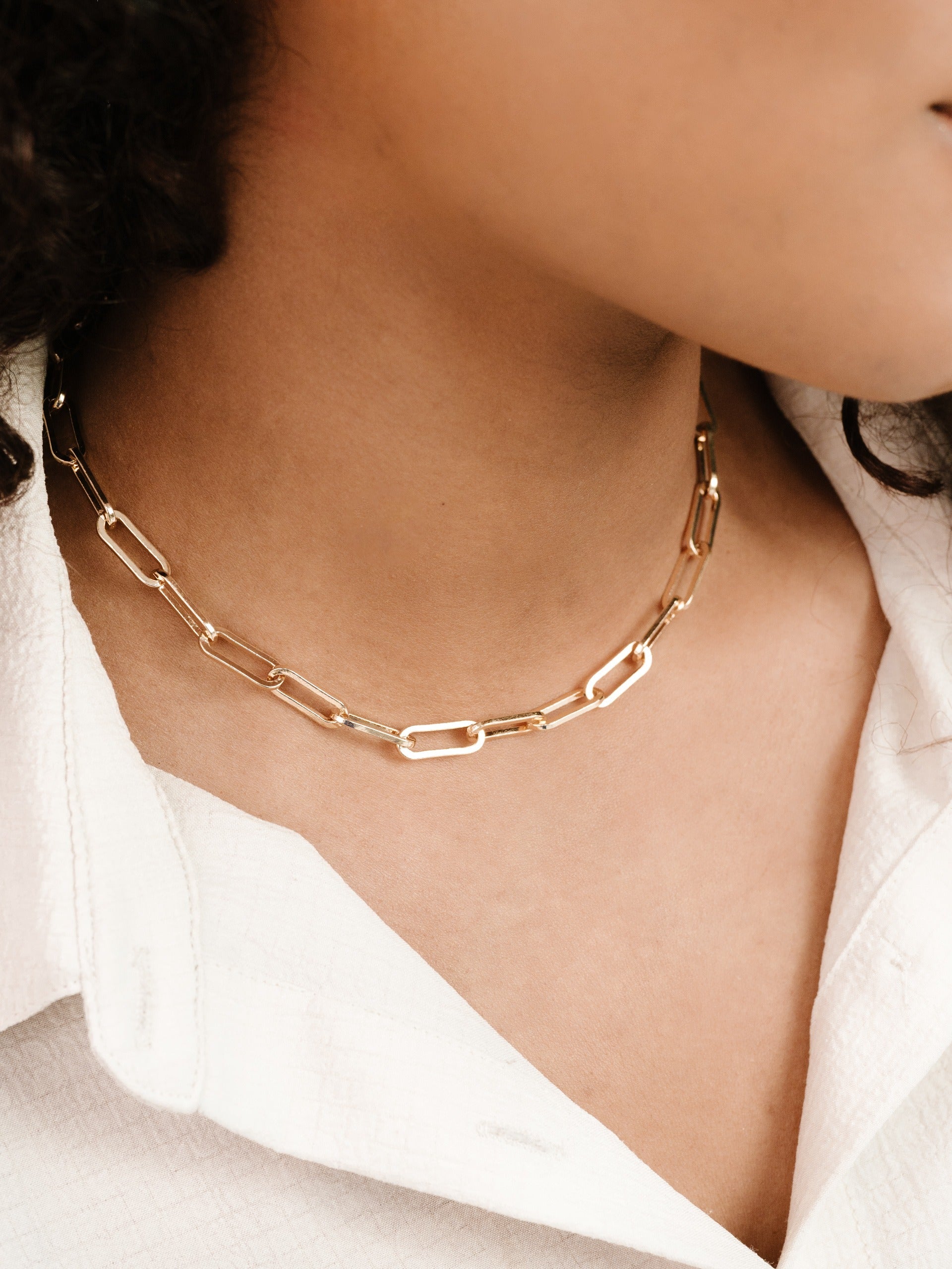 A gold chain necklace with elongated links rests on a model's neck, complementing a white collared shirt. The setting appears casual, focusing on the jewelry's elegant design.