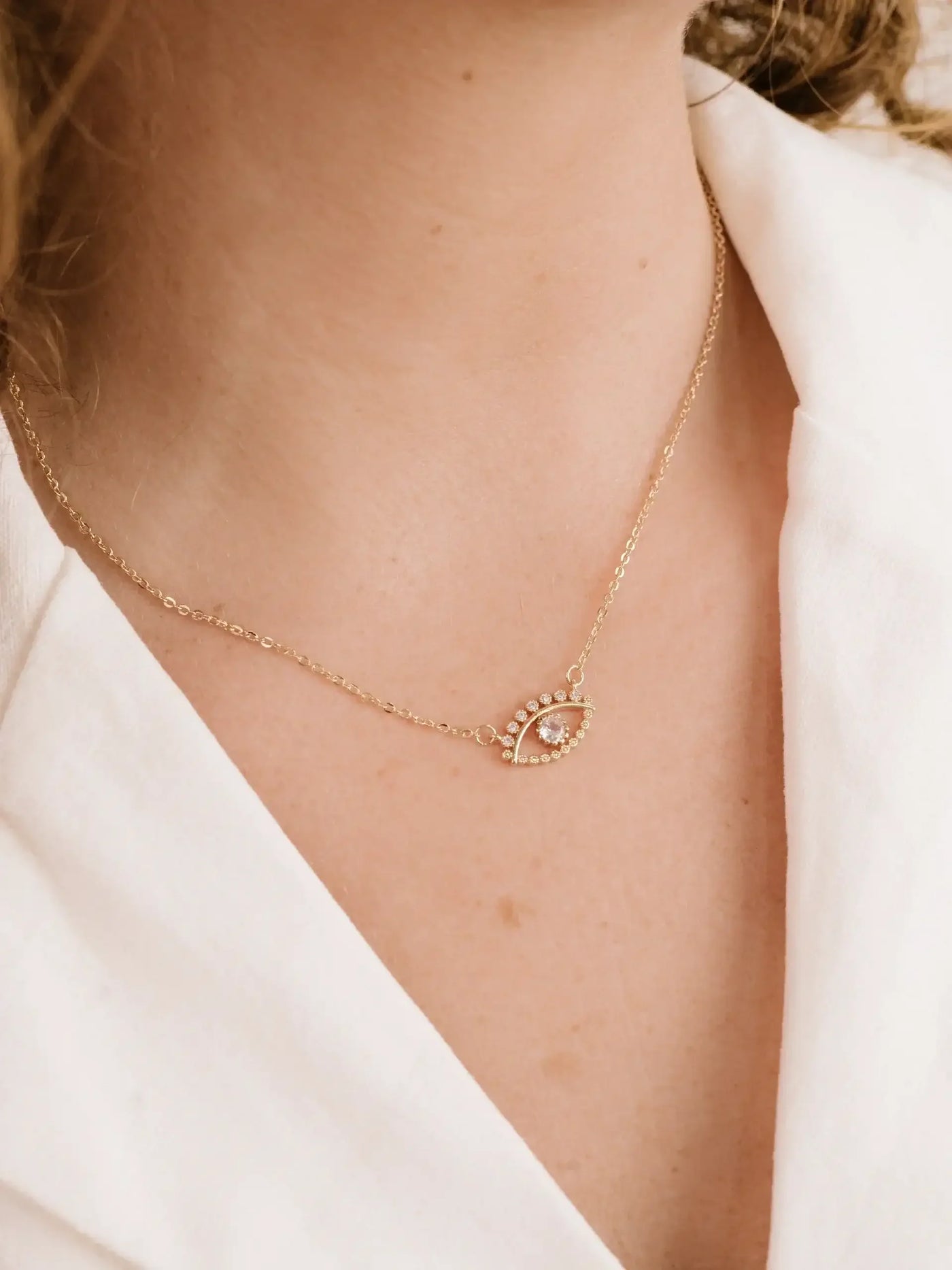 A delicate gold necklace featuring an eye-shaped pendant adorned with small stones rests on the neck of a person, who is wearing a white fabric top, creating a soft and elegant appearance.