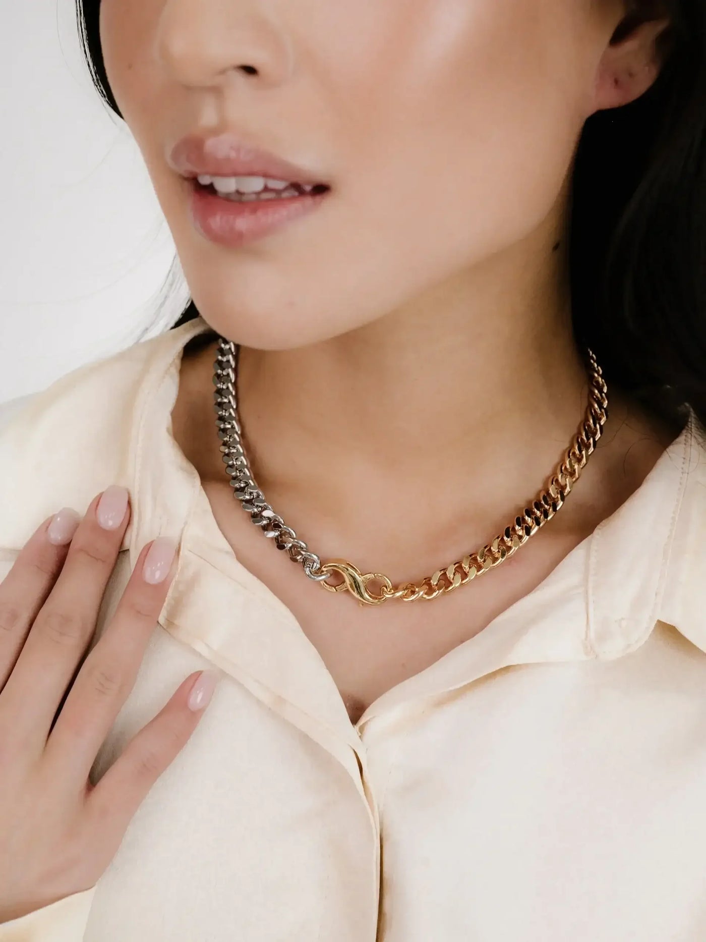 A close-up of a womanâ€™s neck showcases a stylish necklace made of interlocking silver and gold chains. She lightly touches the necklace while wearing a soft, cream-colored blouse.