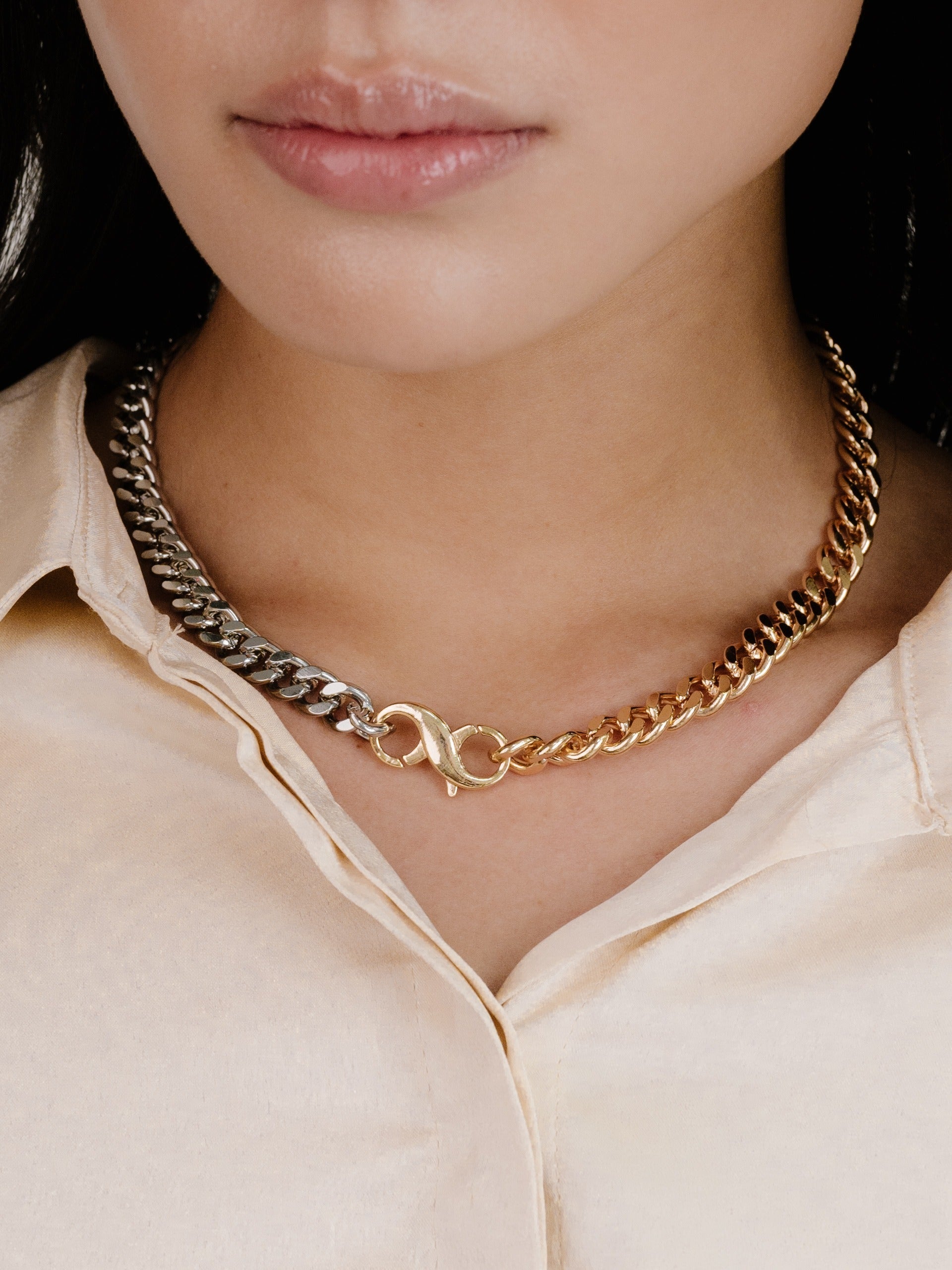 A silver and gold chain necklace with an infinity symbol is worn around the neck of a person, who has smooth lips and is dressed in a light-colored, button-up shirt.