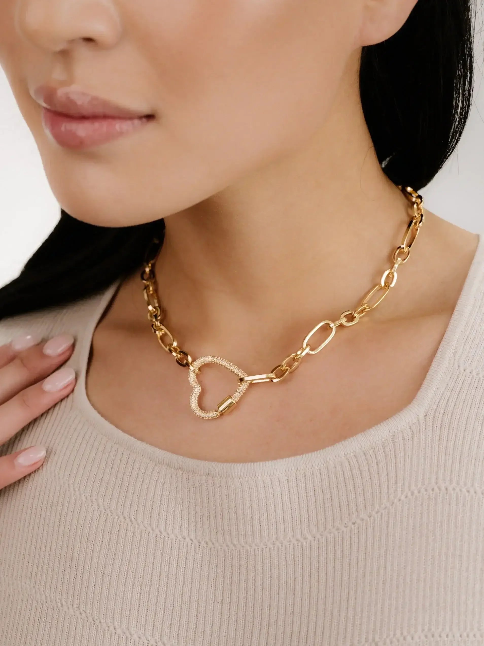 A golden heart-shaped necklace rests on a woman's neck, while she gently touches it with her fingers. She wears a light, textured top, and has straight dark hair.