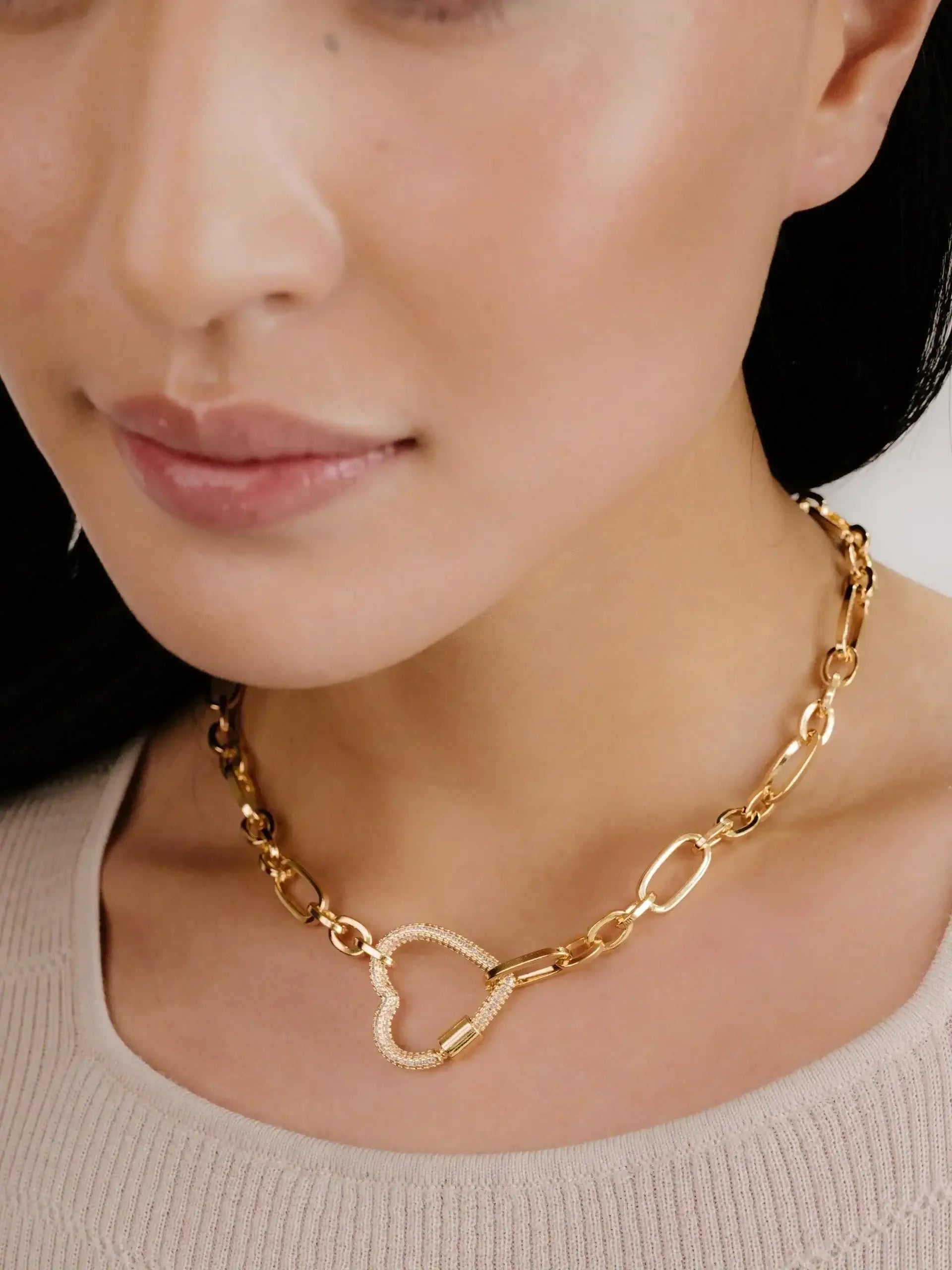 A gold necklace featuring a heart-shaped pendant rests on the neck of a person. The individual has smooth skin and dark hair, wearing a neutral-toned top.