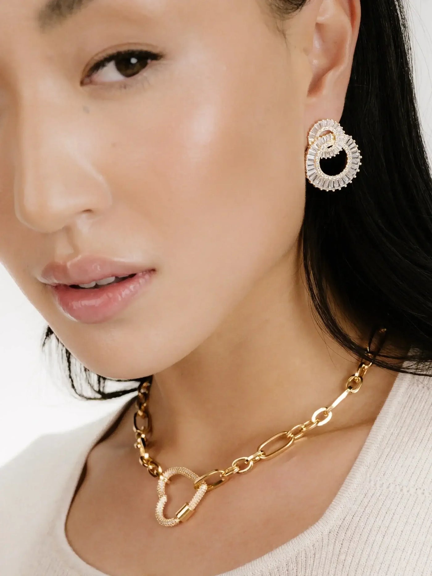 A close-up of a model's face showcases a pair of intricate gold earrings and a gold chain necklace. The background is softly blurred, emphasizing the jewelry and the model's elegant features.