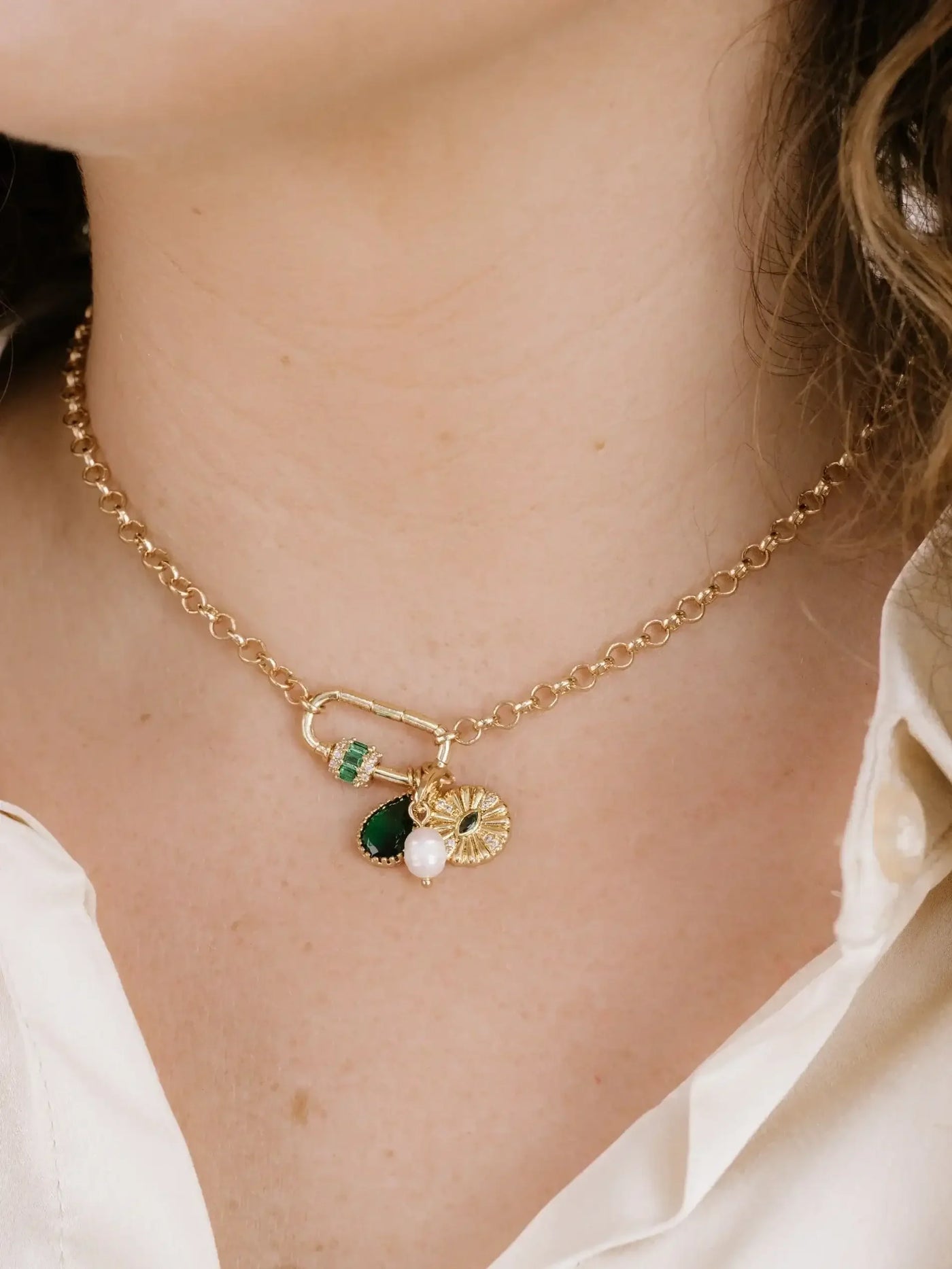 A gold chain necklace adorns the neck, featuring decorative charms like a green gem, a pearl, and a sunflower design. The wearer has light skin and wavy hair.