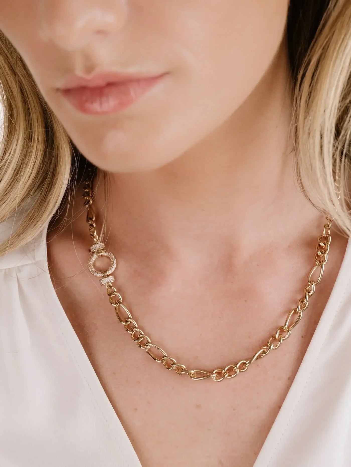 A gold chain necklace with an oval clasp rests on the neck of a woman. The attire features a light-colored, low-cut blouse, emphasizing the necklace against her skin tone.
