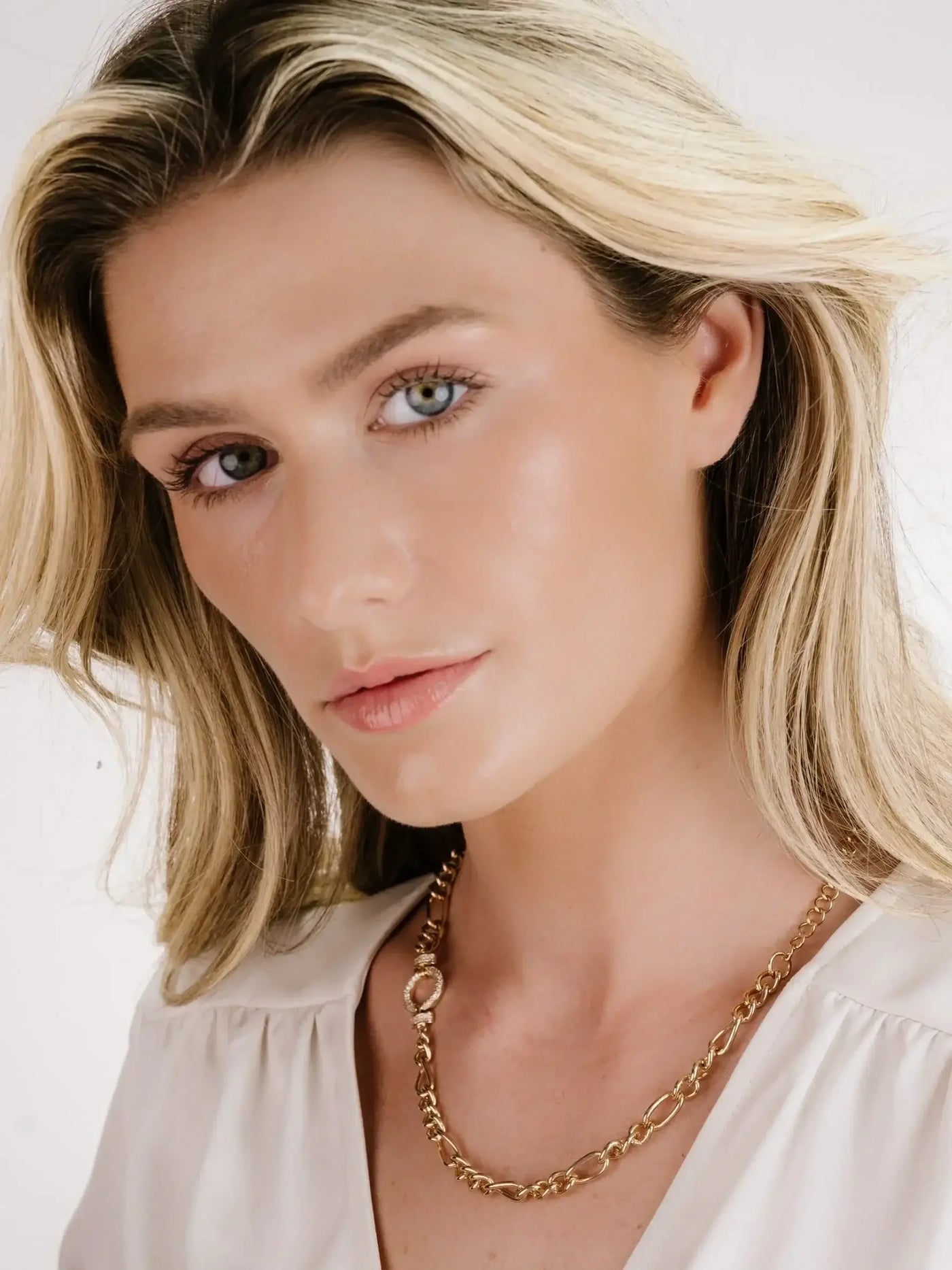 A woman with long, wavy blonde hair poses while wearing a gold chain necklace. She has a natural makeup look, with soft lighting against a neutral background.