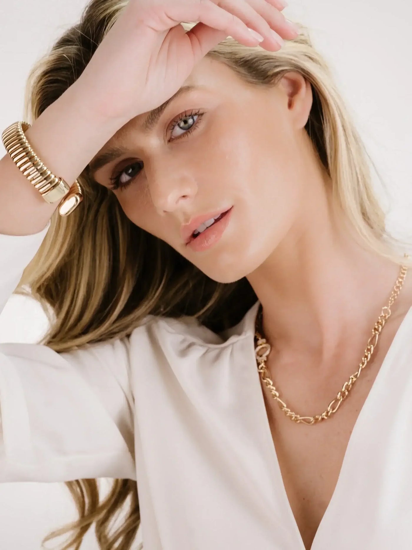 A woman with long, wavy hair poses gently, resting her hand on her forehead. She wears elegant gold jewelry, including a bracelet and necklace, against a soft, neutral background.