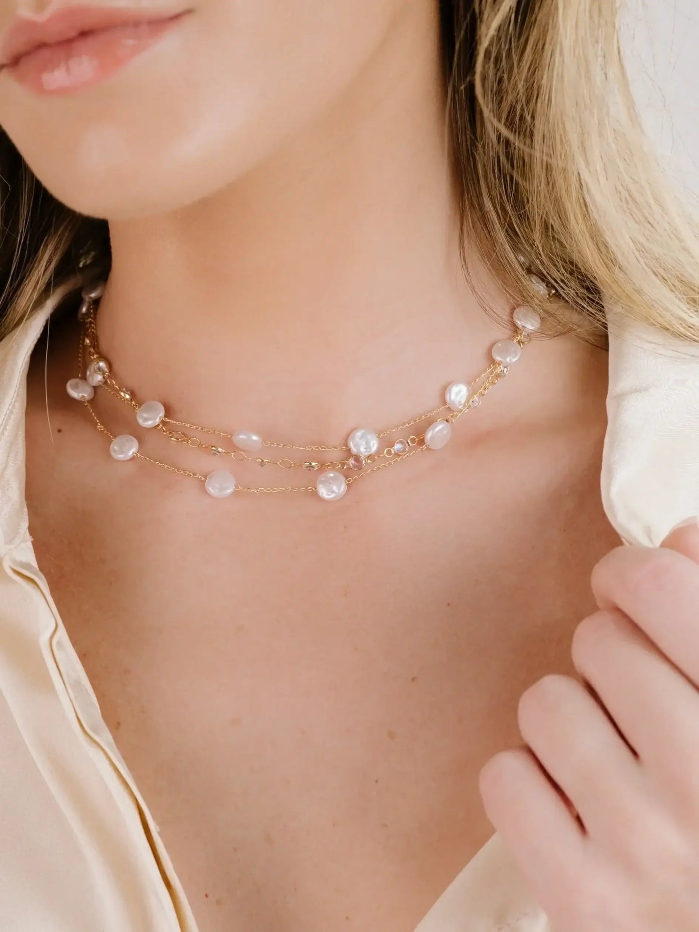 A layered pearl necklace adorns a woman's neck as she lightly touches it, set against a softly lit background that complements her elegant, silky blouse.