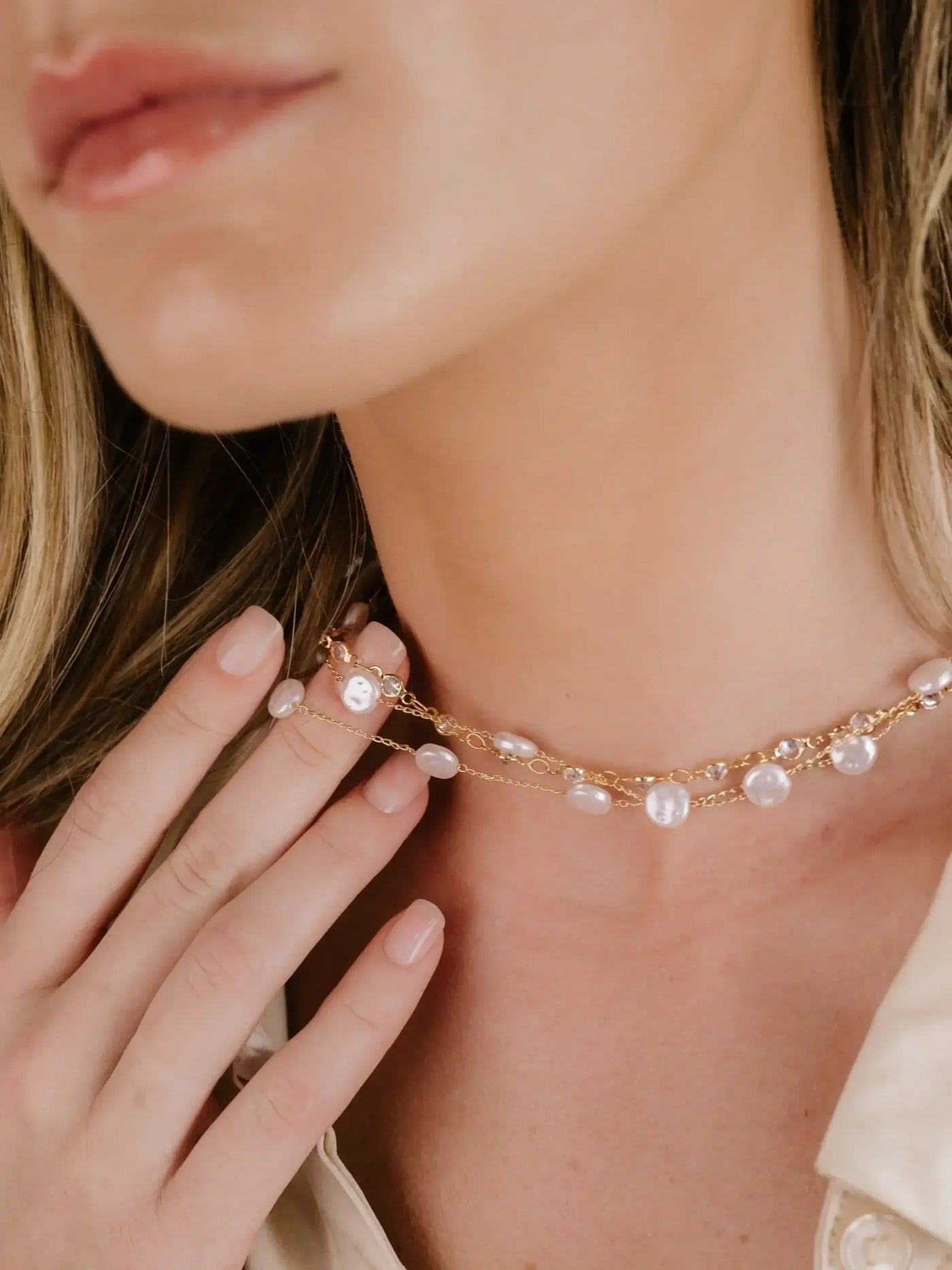 A delicate gold chain necklace with pearl-like beads is being gently touched by a woman's hand. Her skin and partially visible face suggest a soft, elegant aesthetic, set in a neutral-toned context.