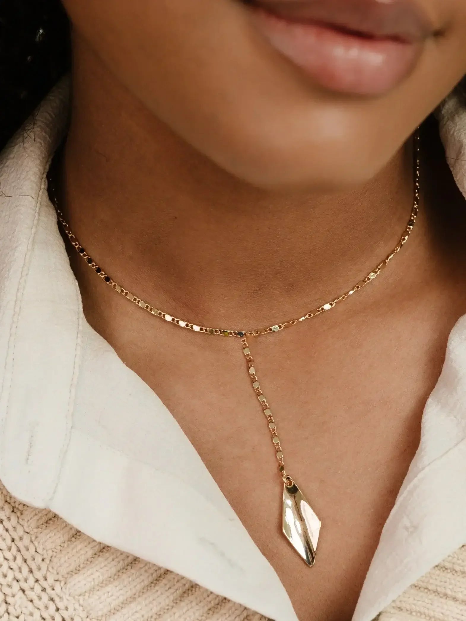 A gold necklace with a decorative, elongated pendant rests on the neckline of a person, whose skin tone is visible against a layered light shirt and a knitted sweater.