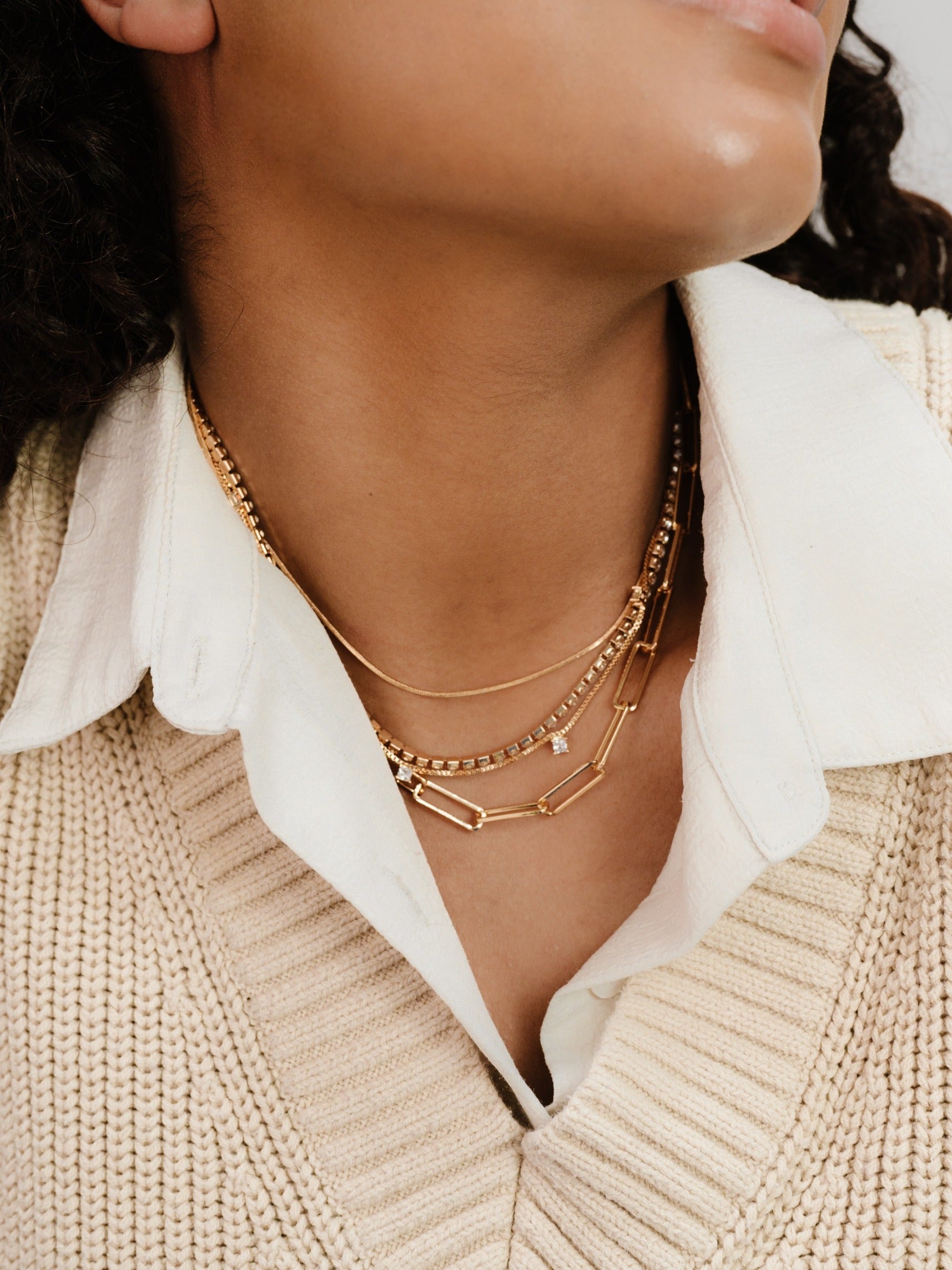 A person's neck adorned with multiple gold necklaces, including chains and one with a small pendant, is partially visible above a light-colored, collared shirt, all set against a neutral background.