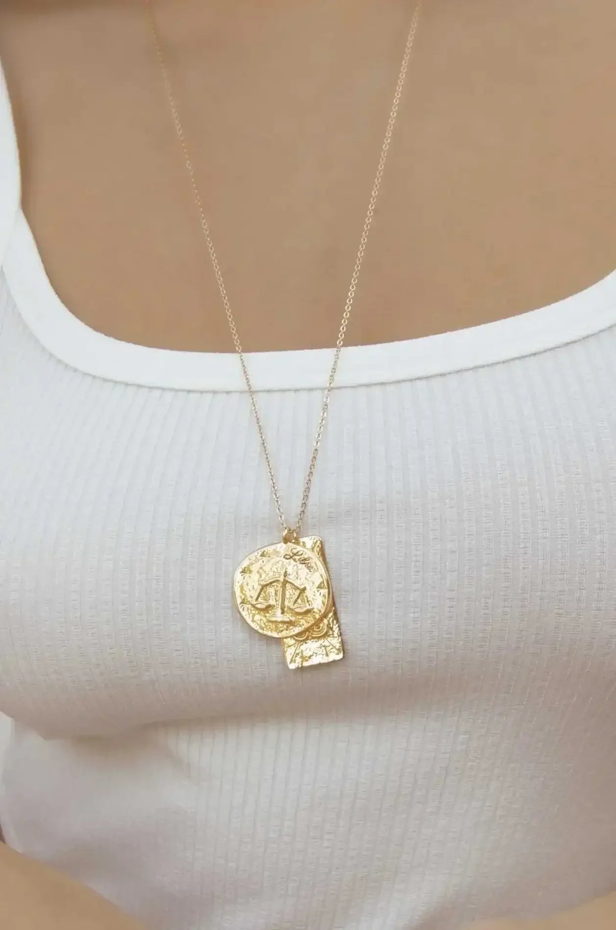 A gold pendant necklace featuring a circular charm with a balance scale design hangs around a person's neck, resting on a ribbed white top, against a neutral background.