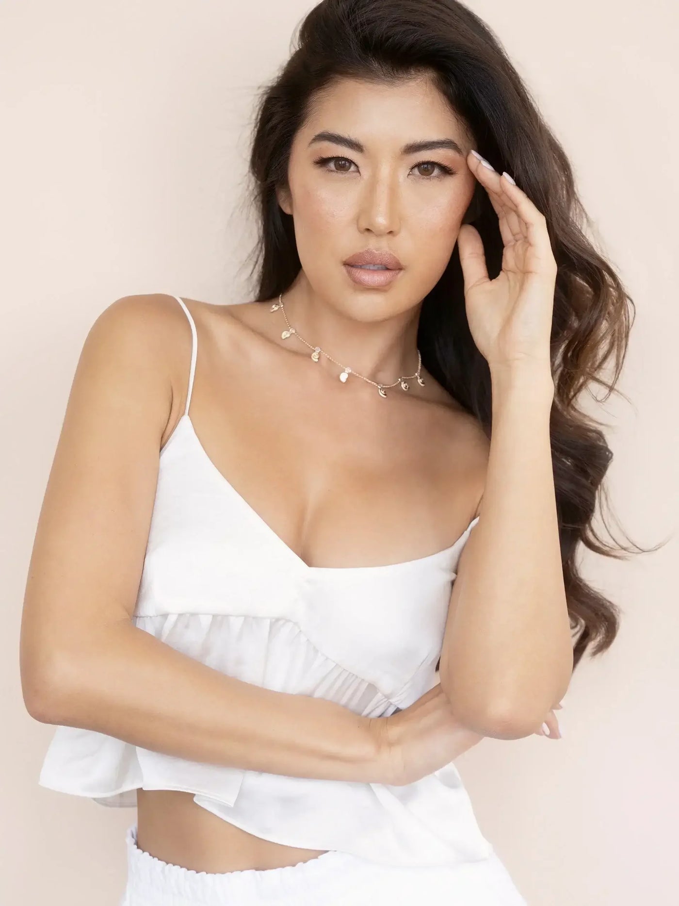 A woman with long, dark hair wearing a white sleeveless top poses elegantly, gently touching her face with one hand in front of a soft beige background.