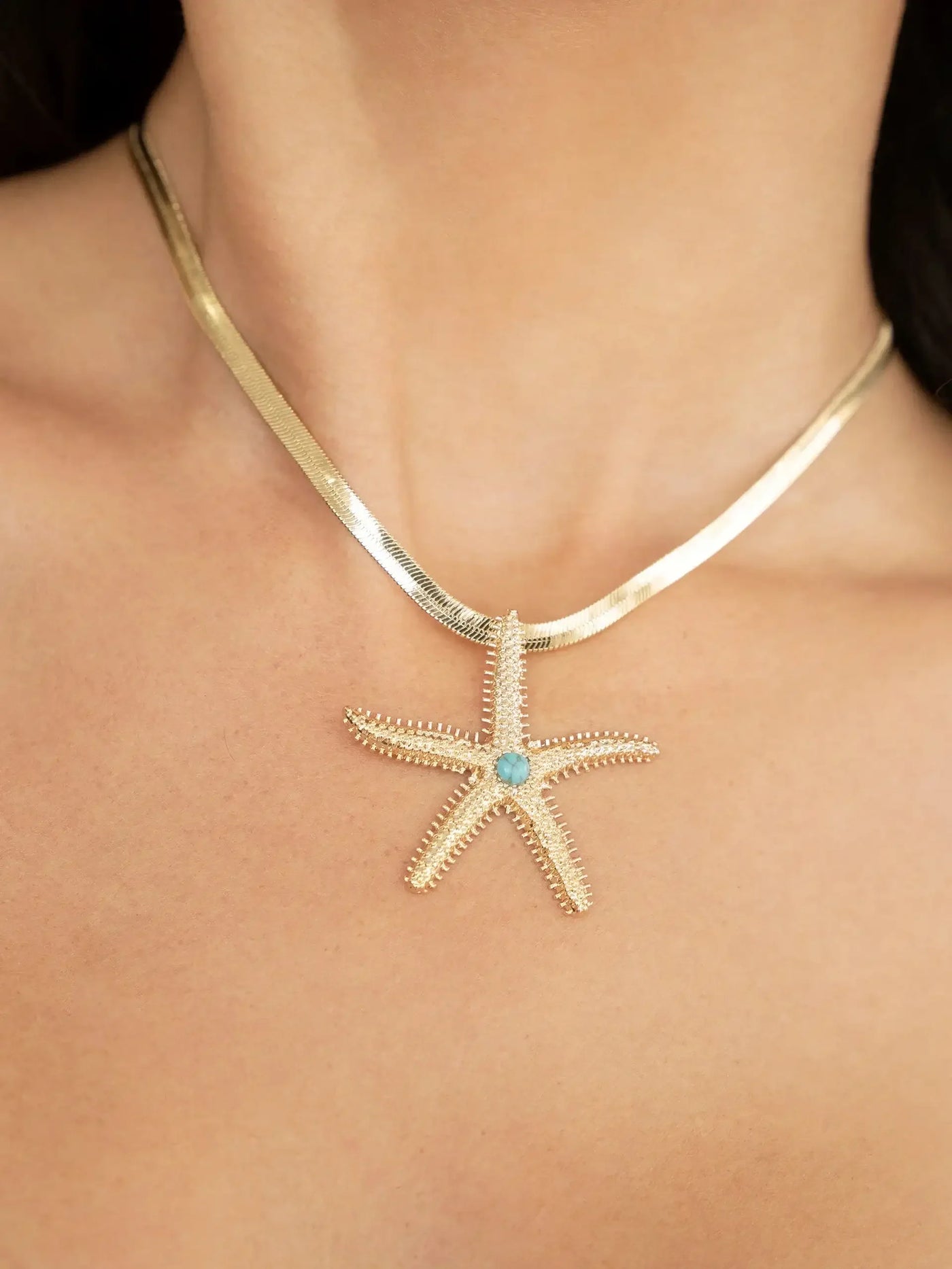 A gold necklace features a starfish pendant with intricate detailing and a blue gemstone at its center. It hangs on a bare neck against a neutral background.