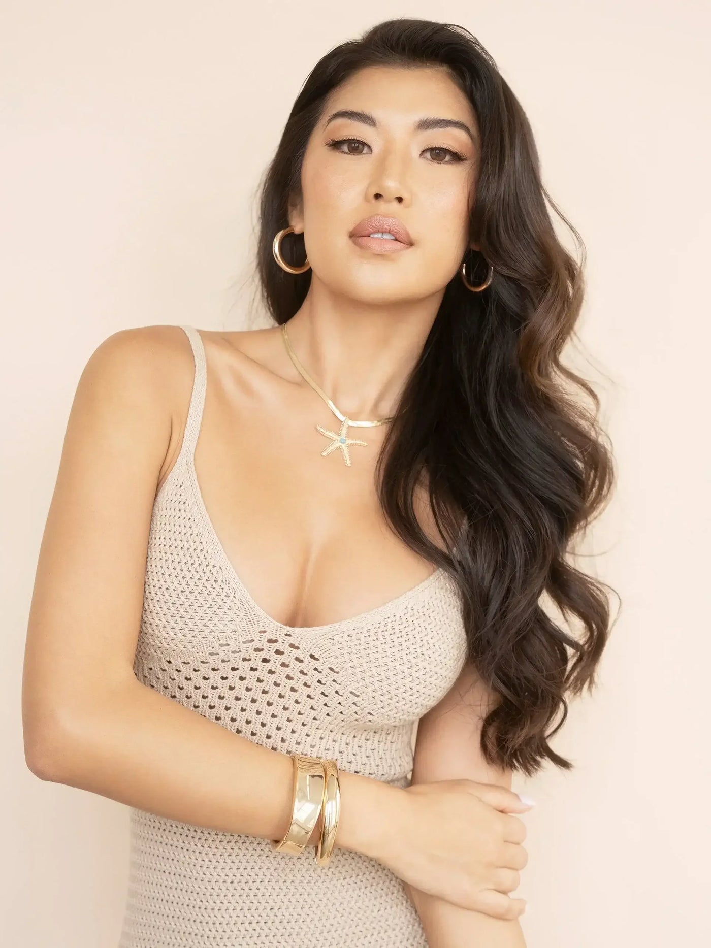 A woman with long, wavy hair poses confidently, wearing a sleeveless beige knit top and gold accessories. The neutral background enhances her sophisticated appearance and focused expression.