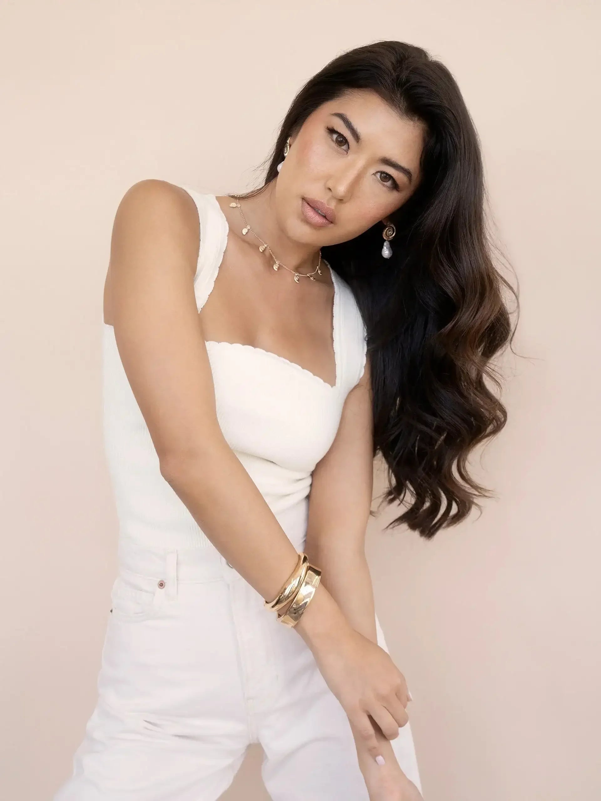 A young woman in a white tank top poses confidently, her long, wavy hair cascading to one side. She wears a delicate necklace and gold bracelets, with a soft, neutral background.