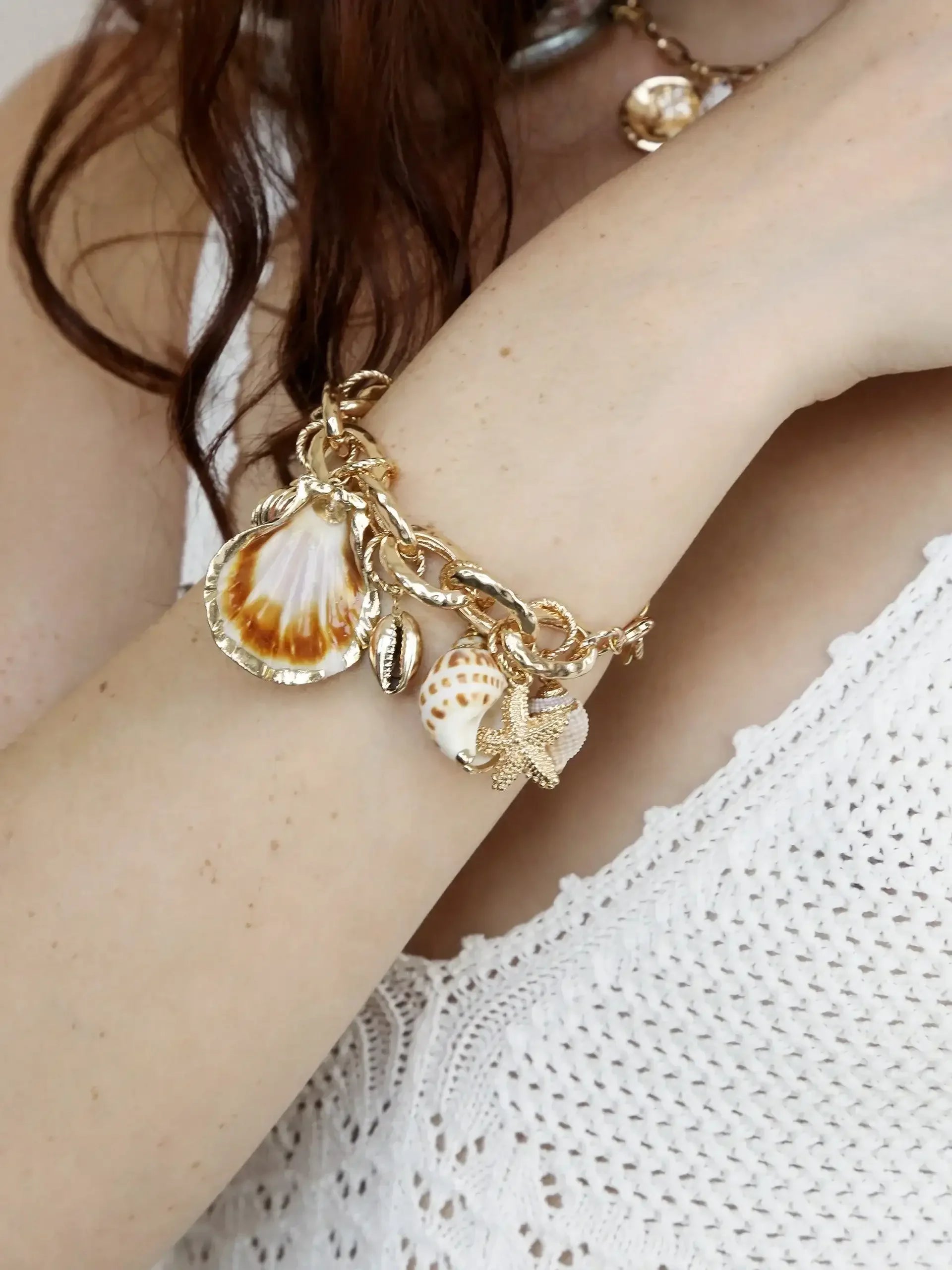 A gold charm bracelet adorned with seashells and a starfish rests on a bare arm. The wrist is partially covered by a knitted white garment, suggesting a casual, coastal aesthetic.