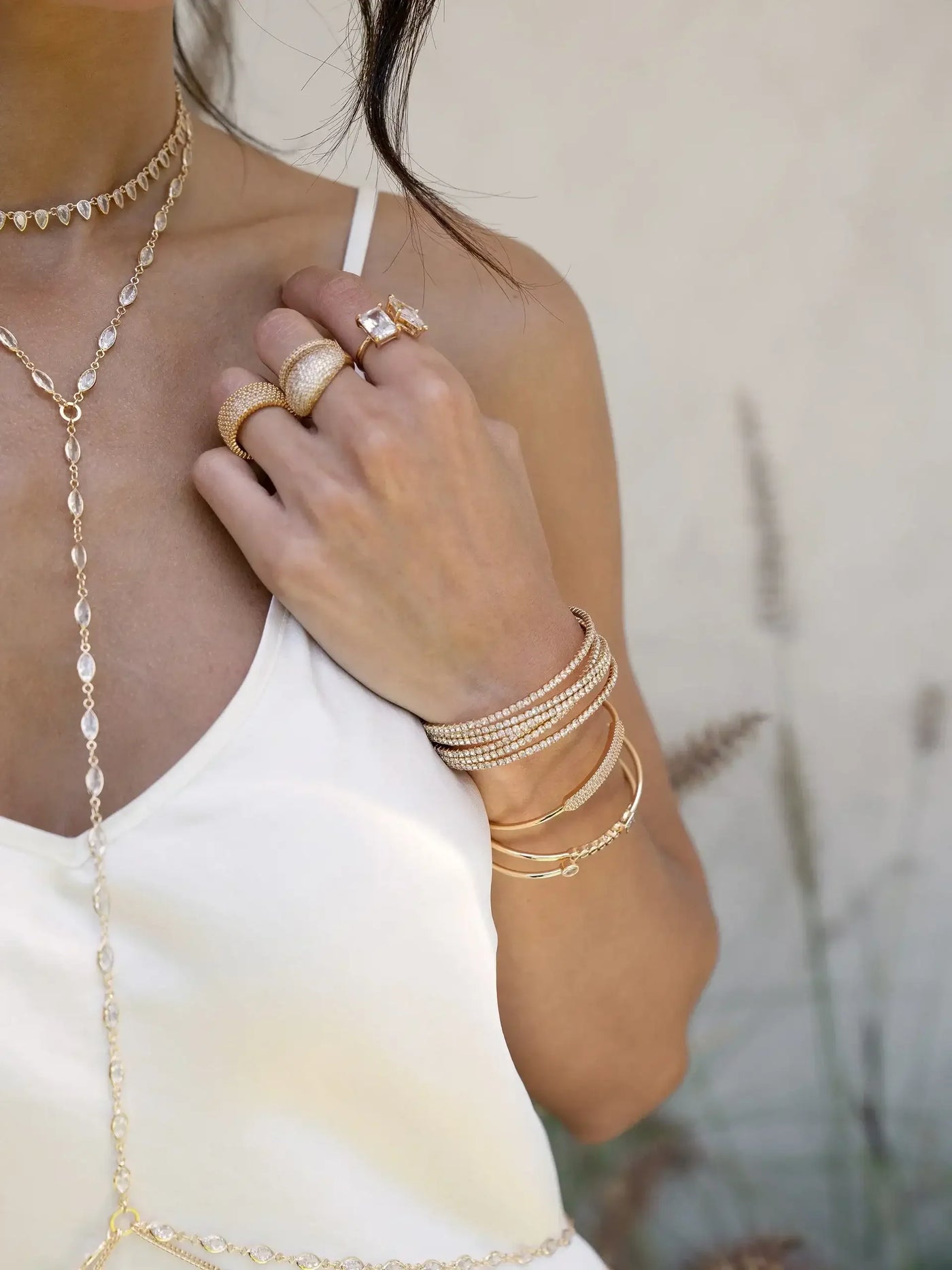 A hand adorned with various gold jewelry, including rings and bracelets, rests on a shoulder, contrasting against a light-colored top and a soft, blurred background of foliage.