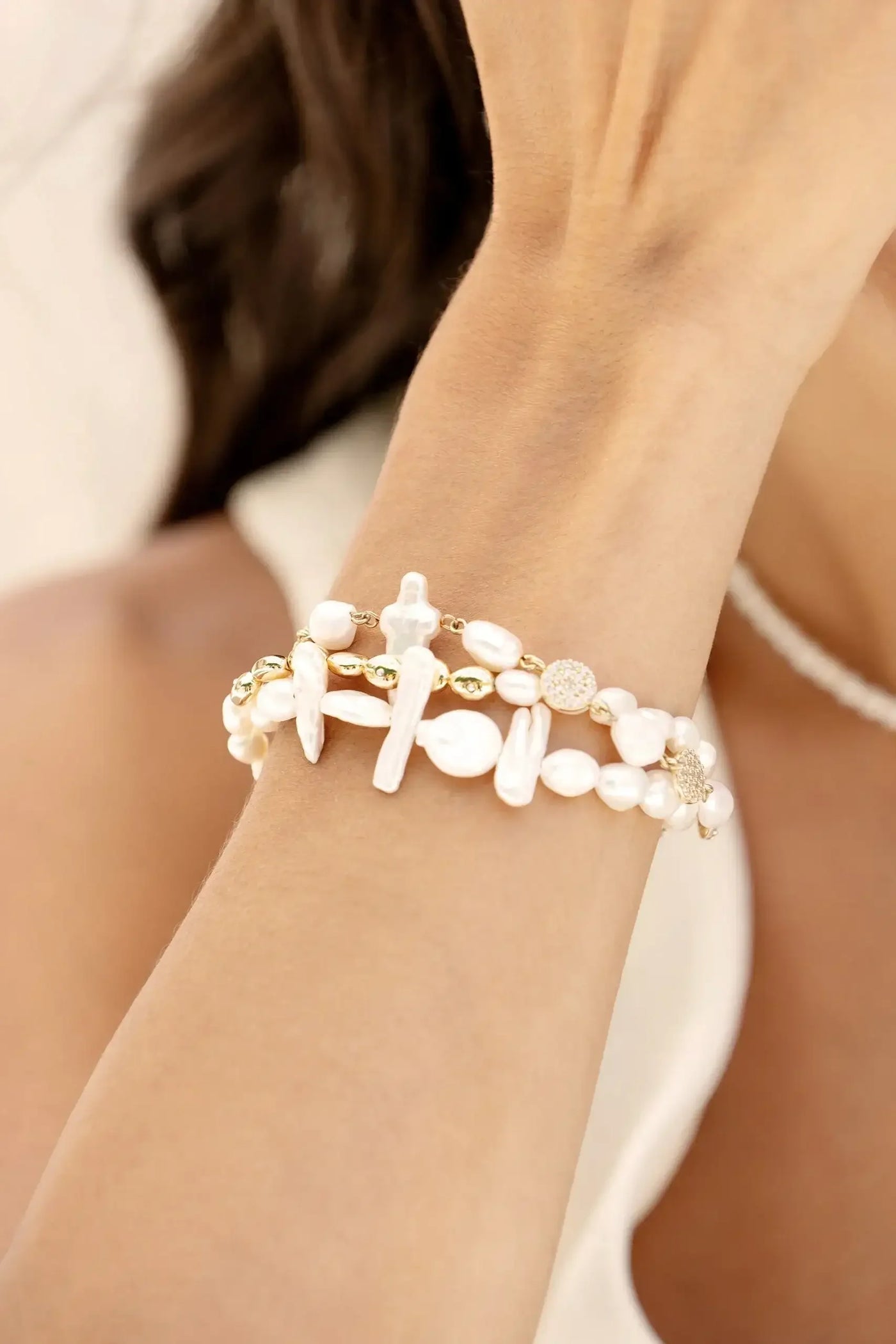 A delicate bracelet made of white pearls and small gold accents adorns a wrist, with the hand raised to touch the face in a softly blurred, light-colored background.