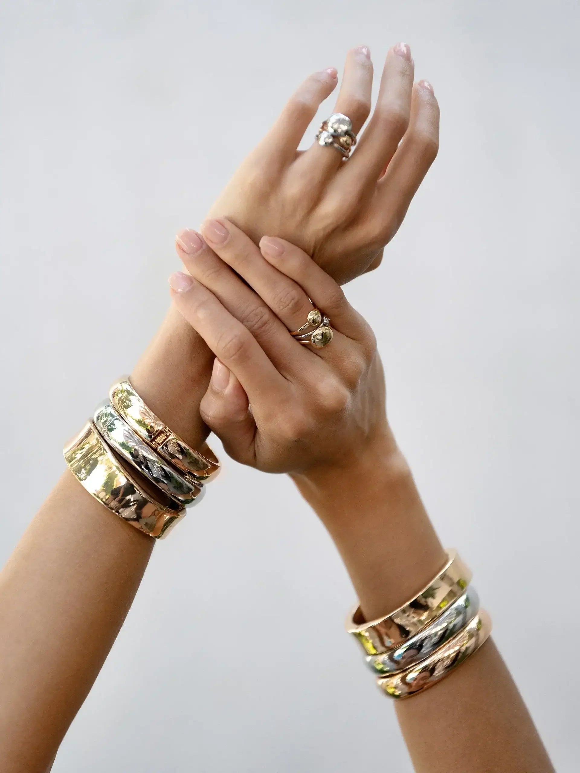 Two hands, adorned with multiple rings and bangles, are clasped together. The fingers have neatly manicured nails, set against a soft, neutral background, emphasizing the elegant jewelry.