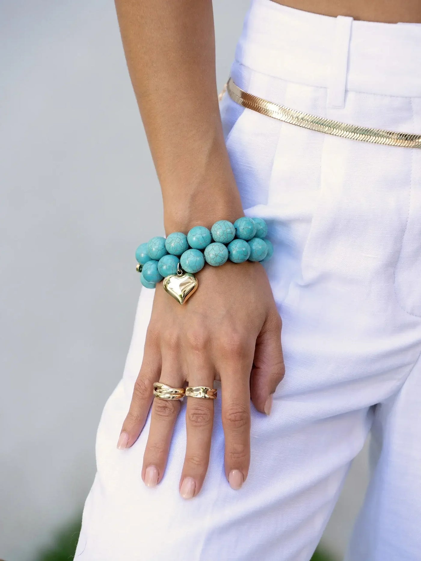 A hand adorned with a turquoise beaded bracelet featuring a gold heart charm, while wearing two gold rings. The context includes a light-colored outfit set against a soft background.