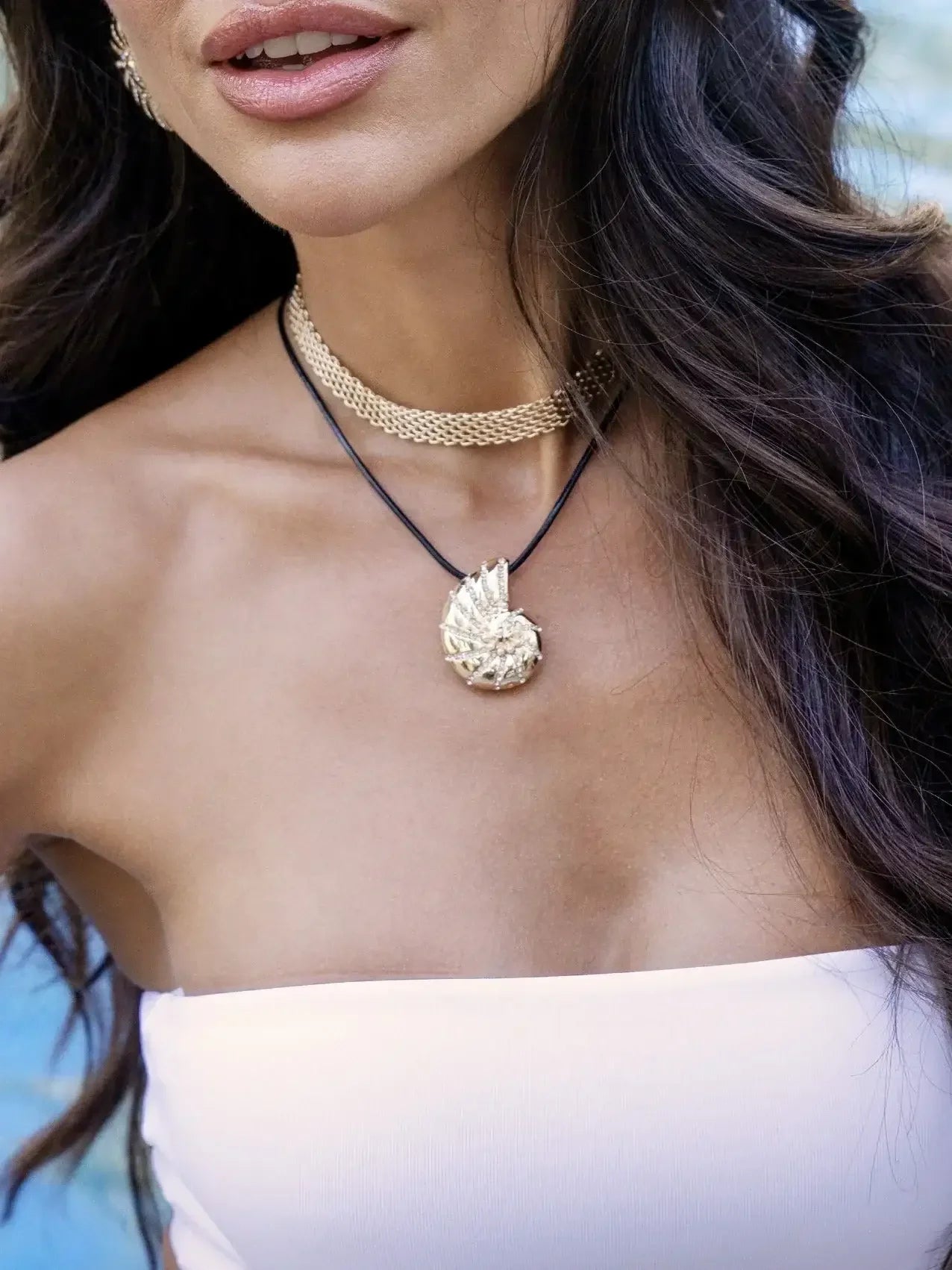 A womanâ€™s neck features two necklaces: a gold chain and a pendant necklace, with a textured, leaf-like pendant. Her long hair falls over her shoulder in a natural setting.