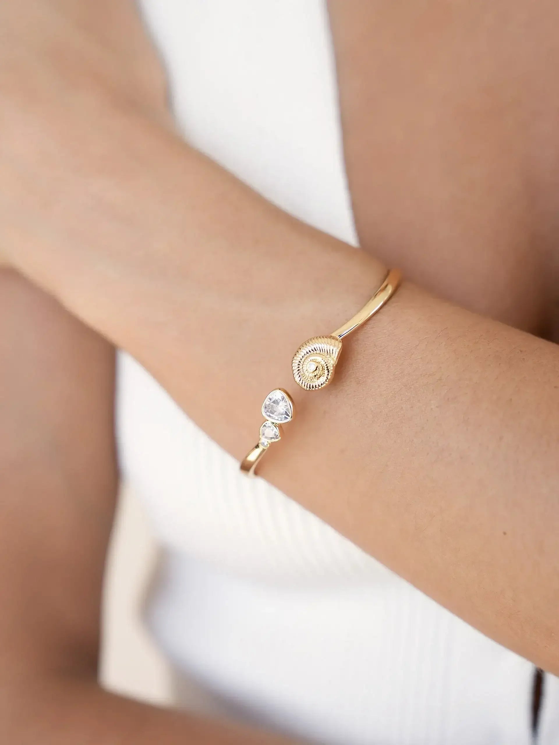 A gold bracelet rests on a person's wrist, featuring decorative elements like a spiral design and gemstones. The skin tone contrasts with the bracelet, set against a soft, neutral background.