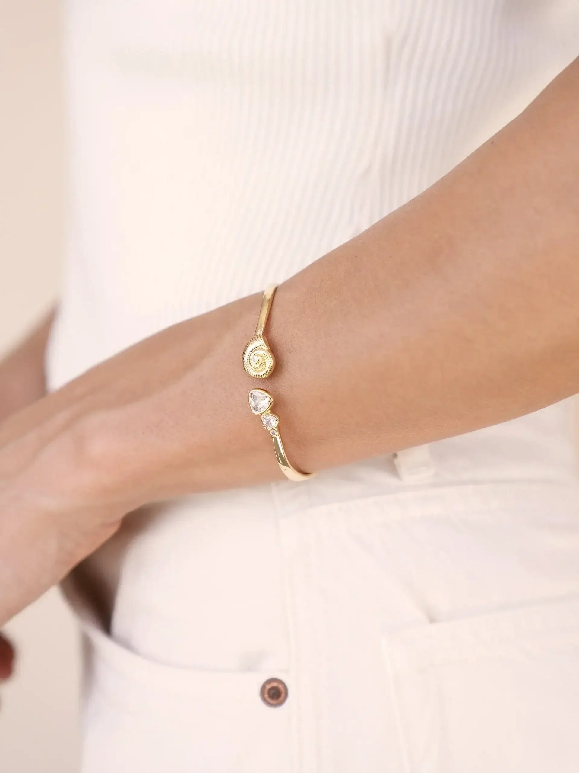 A gold bracelet with a spiral design and two small gemstones is worn on a wrist. The surrounding context features a light-colored, textured top and light-colored pants, suggesting a casual outfit.