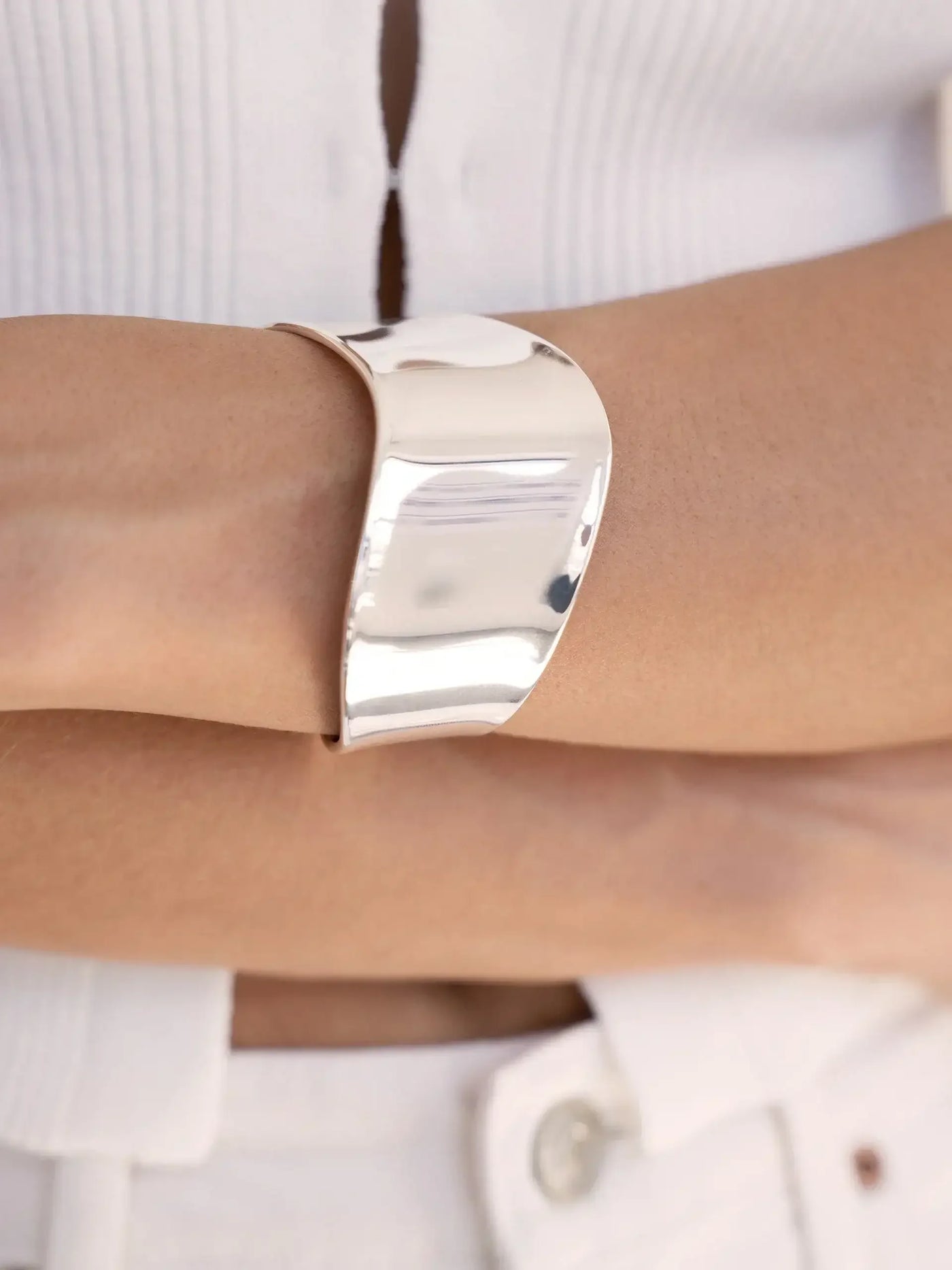 A shiny silver cuff bracelet rests on a person's wrist, which is crossed over the other arm. The individual wears a white, textured top and light-colored pants, suggesting a casual setting.
