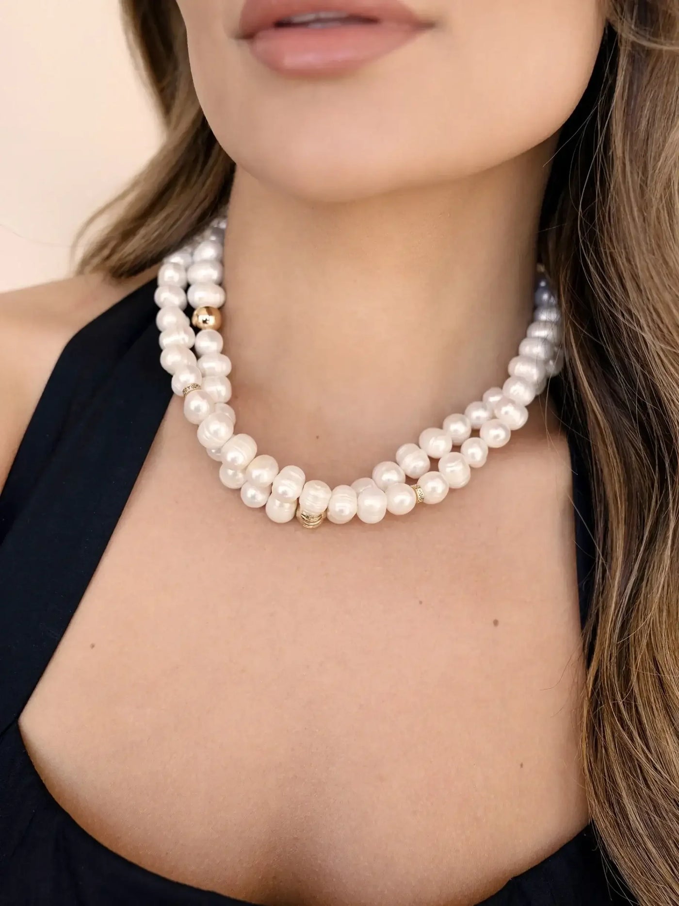 A multi-strand pearl necklace adorns the neck, featuring large white pearls interspersed with smaller golden accents. The wearer is dressed in a dark top, with soft lighting highlighting the skin tone.
