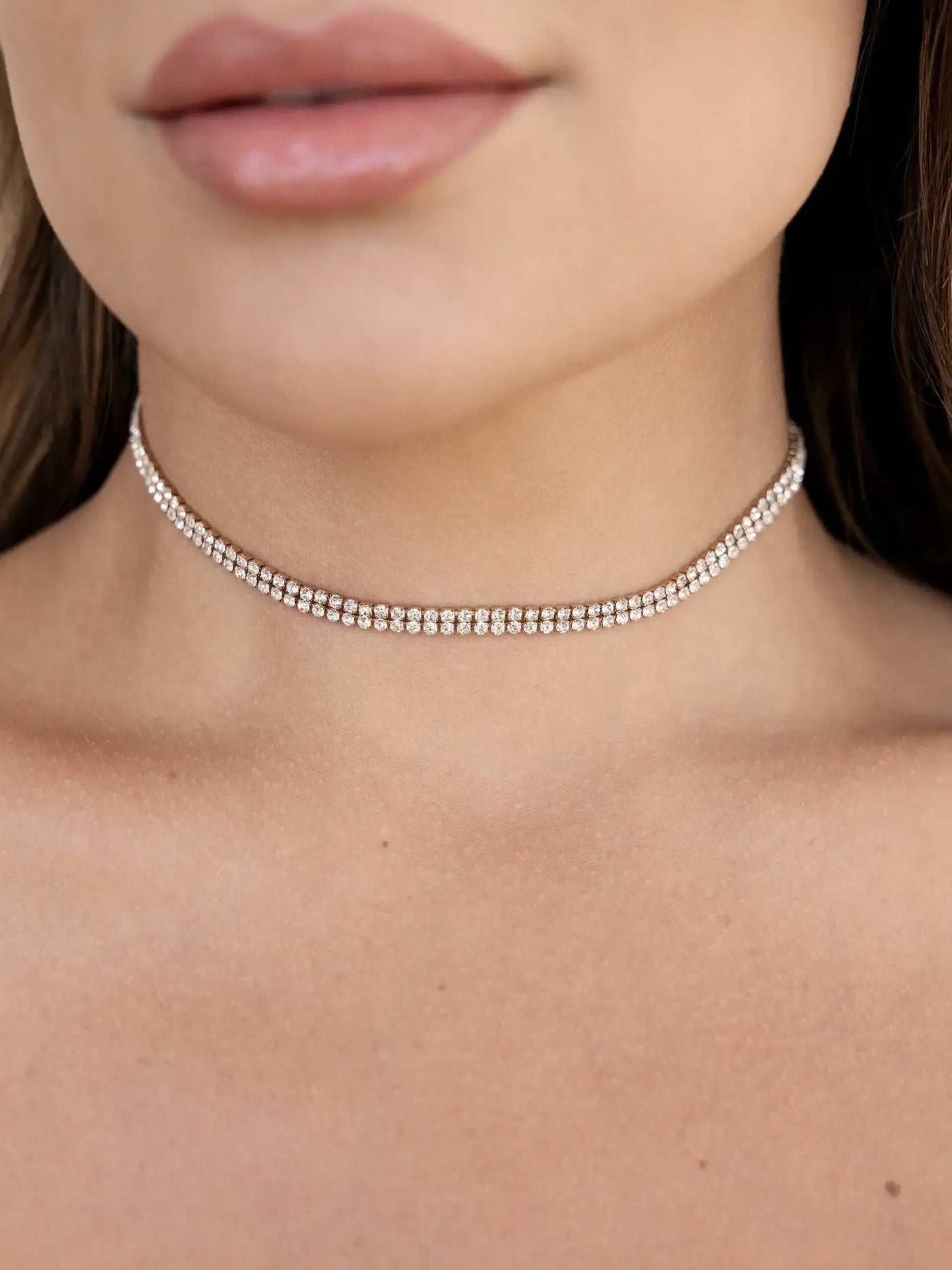 A close-up of a woman's neck showcases a sparkly choker made of small, shiny stones. Her lips appear soft and natural, against a smooth, lightly tanned skin background.