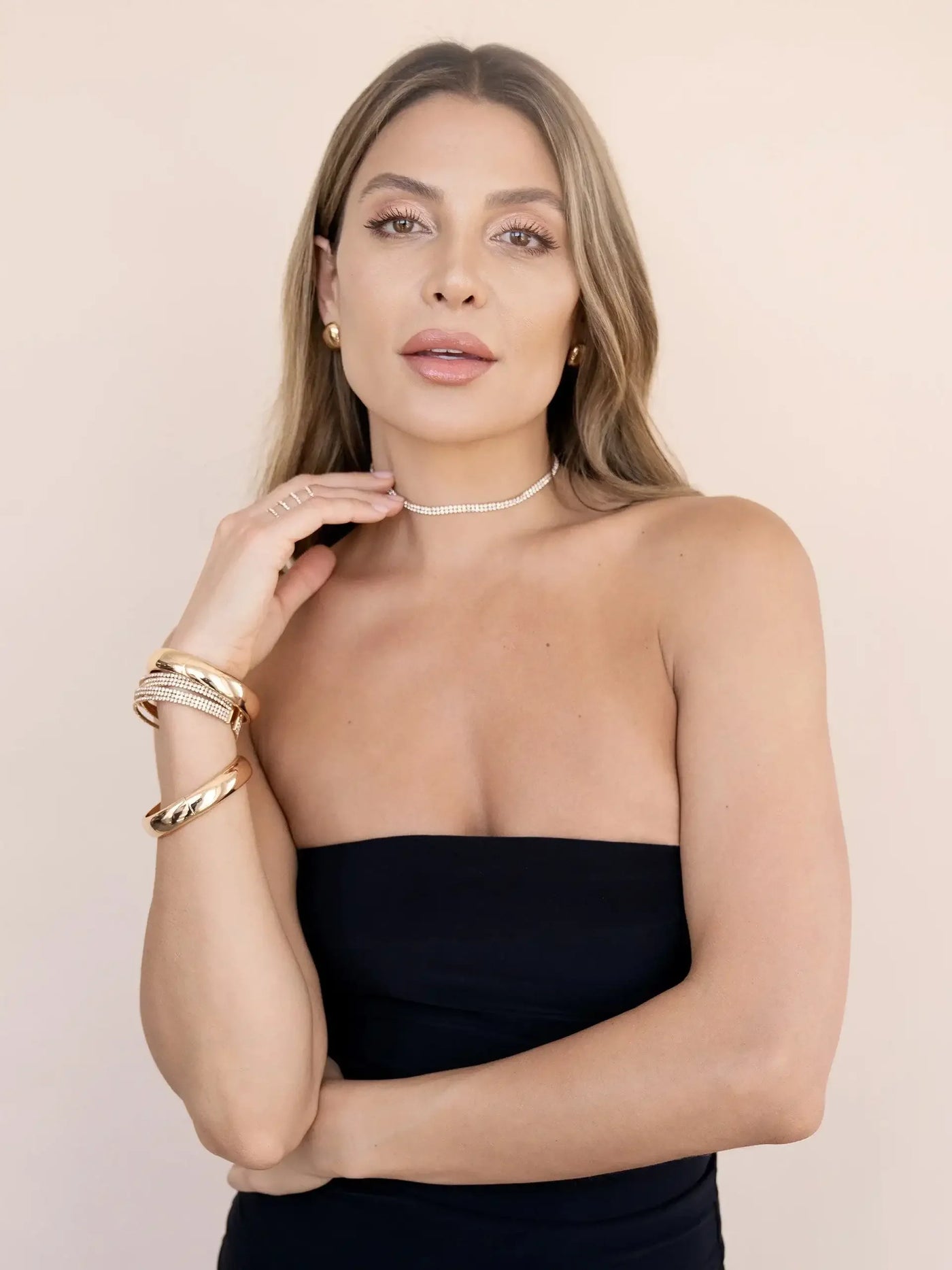 A woman wears a strapless black top, gently placing her hand on a sparkling choker necklace. She displays several gold bracelets, against a soft, neutral background.