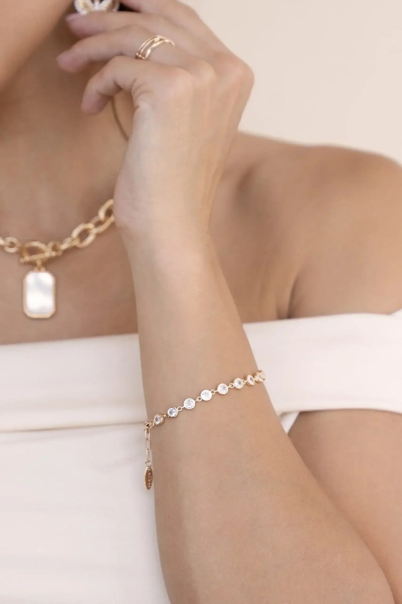 A hand adorned with a delicate gold bracelet featuring circular stones adjusts a pendant necklace. The subject is wearing a strapless top and is set against a neutral background.