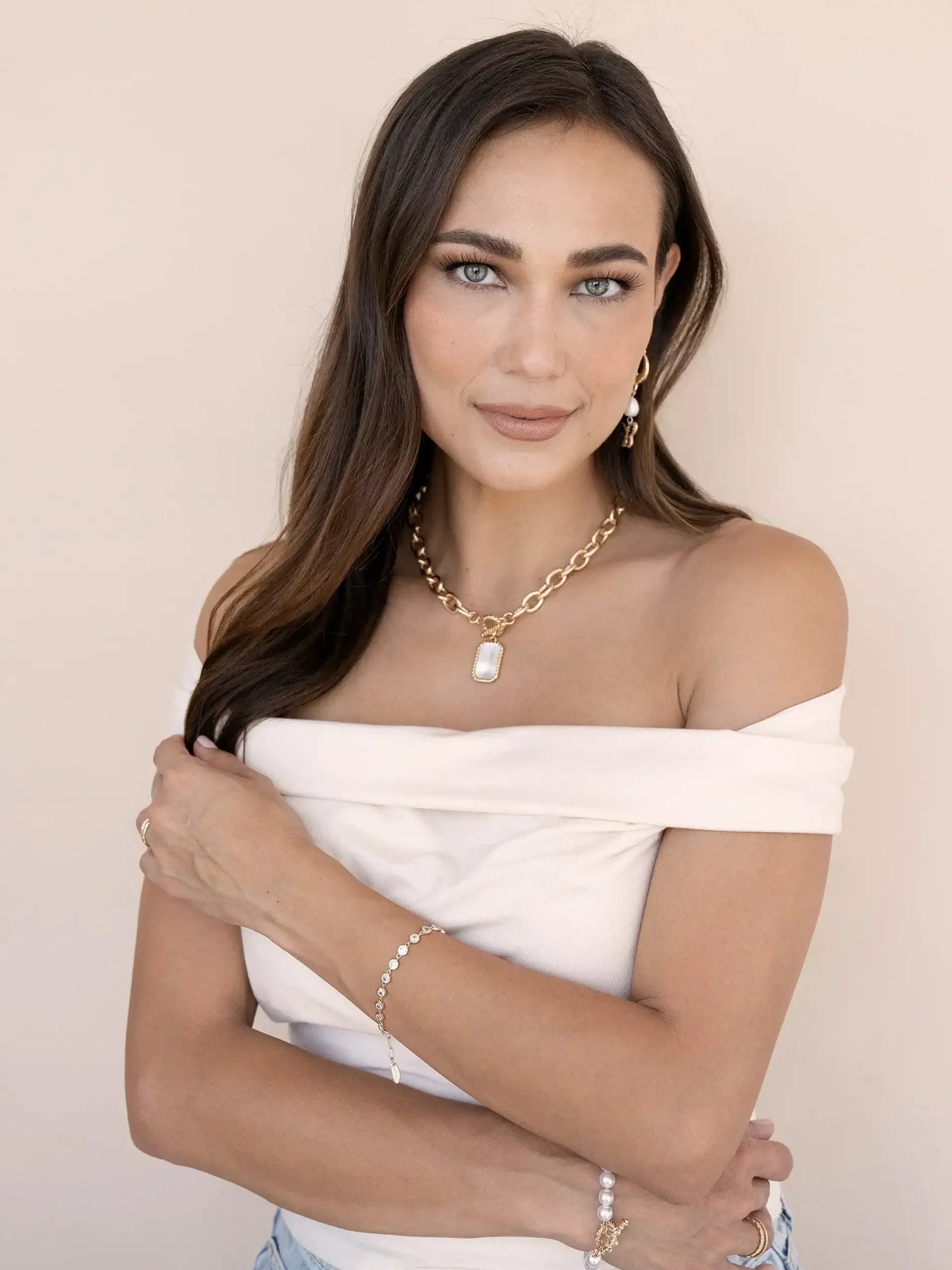 A woman with long brown hair poses gracefully, wearing an off-shoulder top and layered jewelry. She brings her arms around her torso, set against a soft beige background.