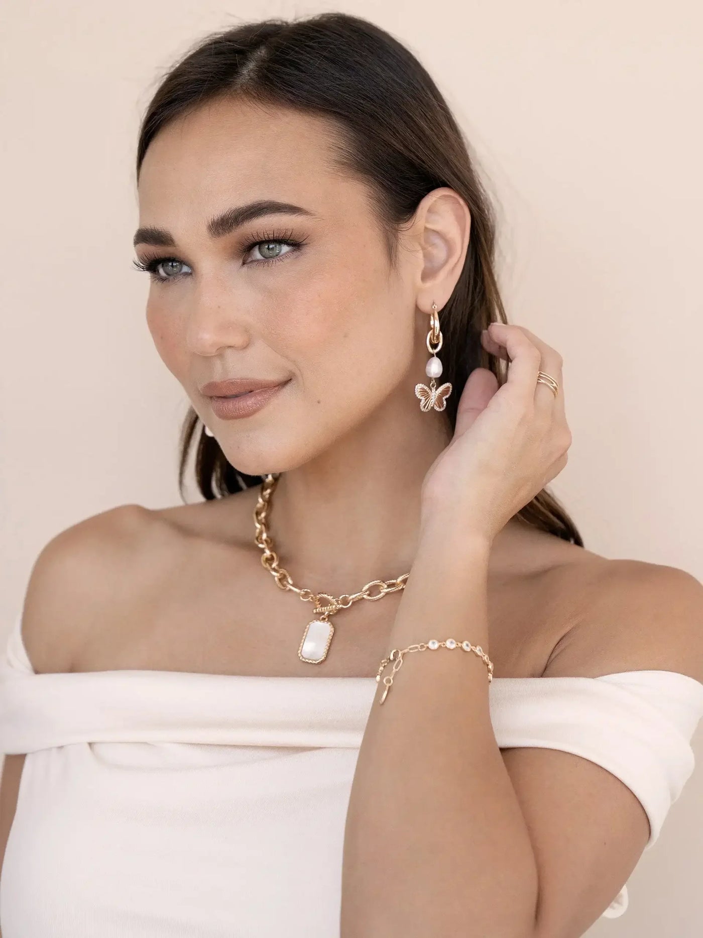 A woman with long dark hair wears elegant jewelry, including a butterfly earring and a chain necklace. She poses gracefully against a soft, neutral background, lightly touching her neck.
