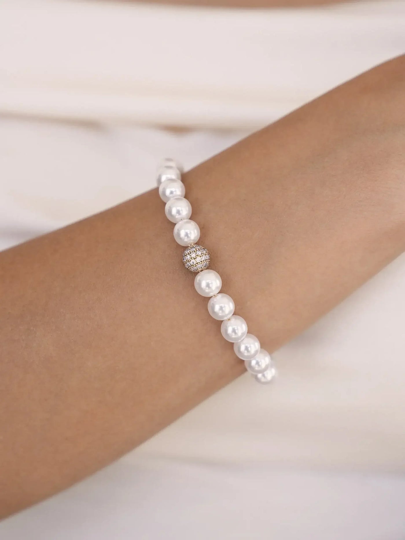 A pearl bracelet adorns a tanned wrist, showcasing alternating white pearls and a decorative gold-accented bead. The background features soft, neutral fabric, enhancing the elegance of the jewelry.