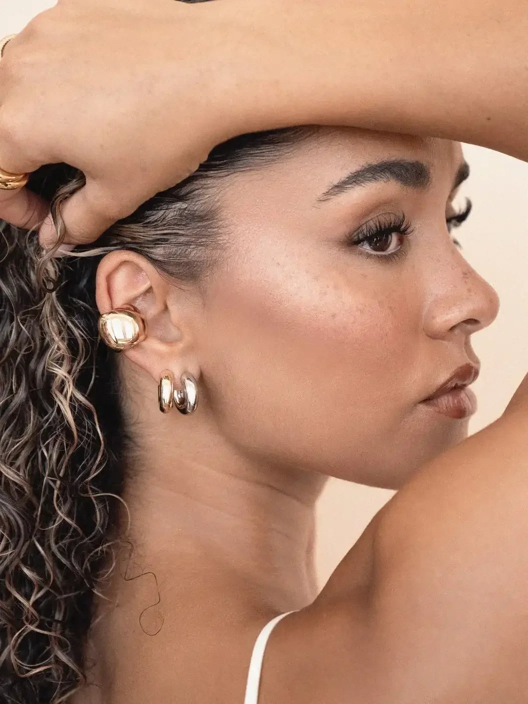A woman with curly hair poses while touching her hair. She wears bold gold earrings and has a subtle, natural makeup look, set against a neutral background.