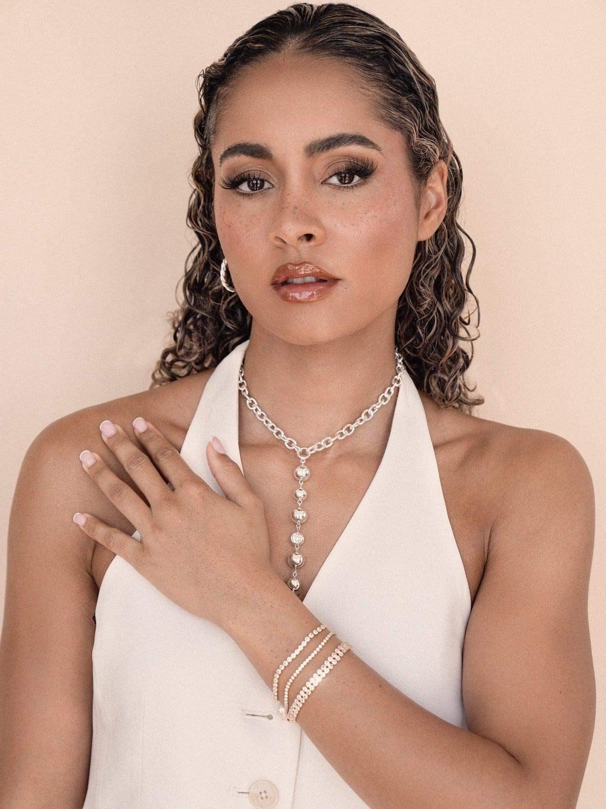 A woman with curly hair wears a white halter top and holds her hand on her chest, showcasing silver jewelry, against a soft, neutral background.