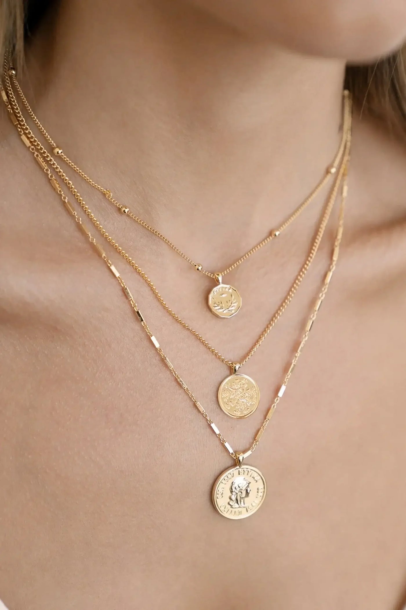 Three layered gold necklaces adorn a person's neck, featuring intricate coin pendants. The skin tone is visible, and the soft lighting enhances the jewelry's shine against a neutral background.