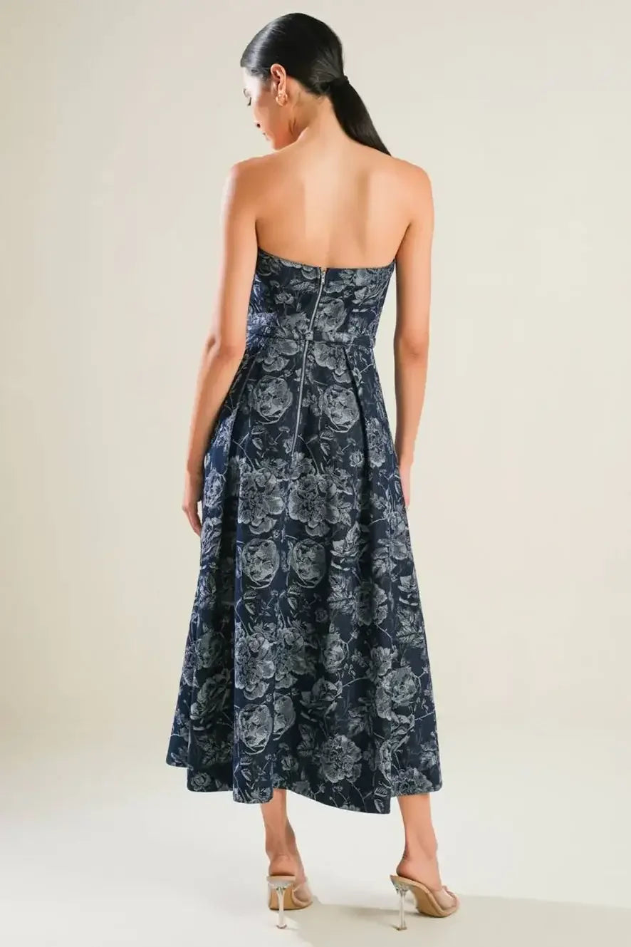A woman stands wearing a strapless, floral-patterned navy dress that flows down to her ankles. She faces away from the viewer, with a light background enhancing the elegant look.