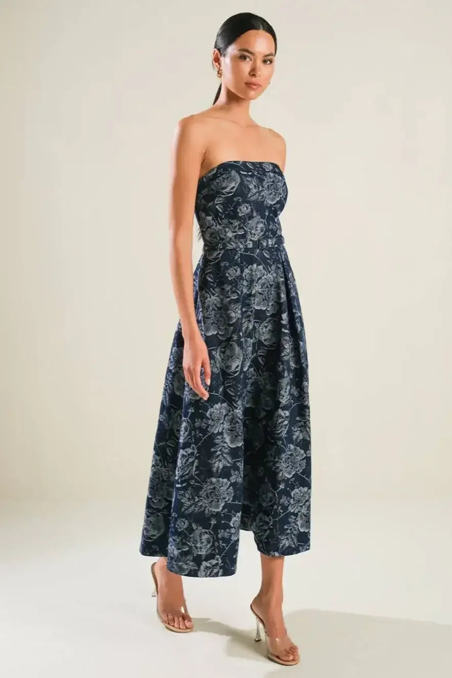 A woman poses gracefully in a strapless, floral-patterned navy dress. She stands on a light-colored floor, showcasing elegance against a simple, neutral background.