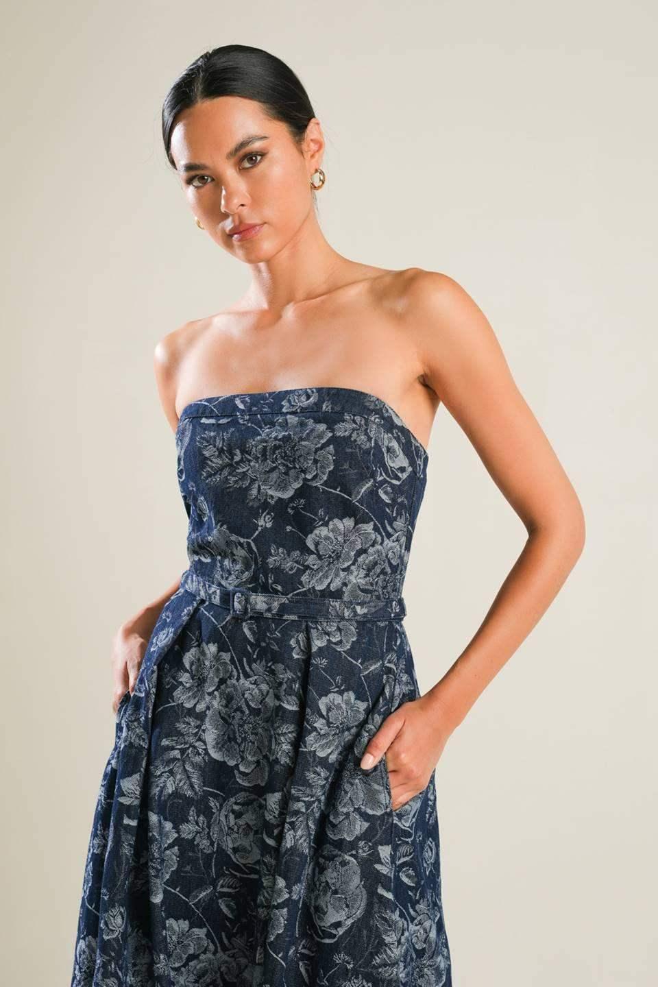 A woman in a strapless, floral-patterned navy dress stands confidently with her hands in pockets. The background is a light, neutral color, highlighting the elegance of her outfit.