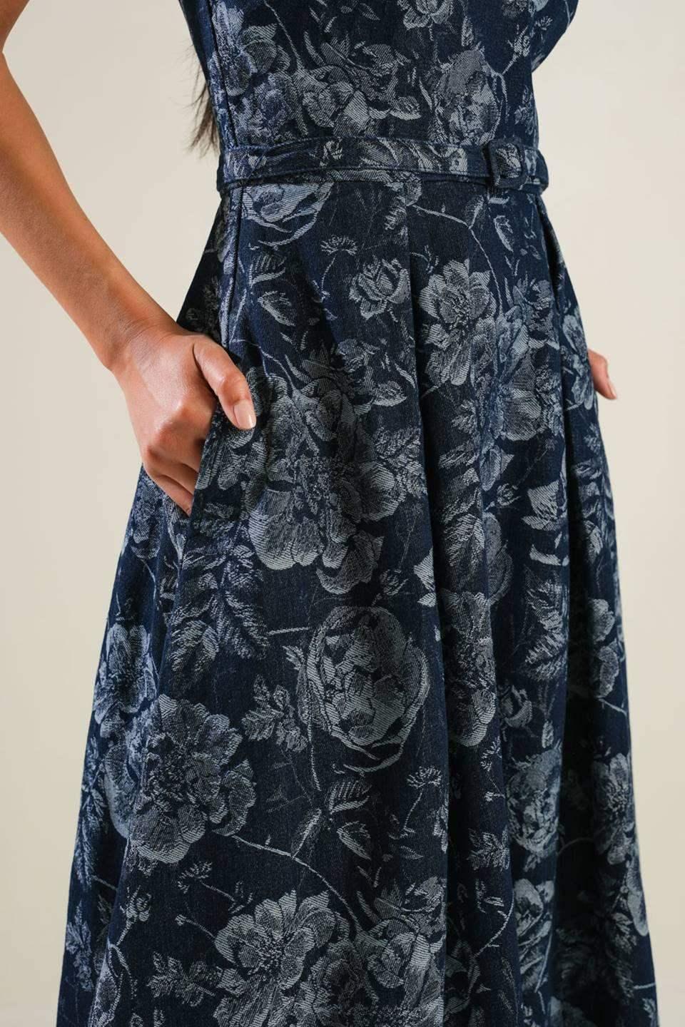 A dark floral dress features a fitted waist and flared skirt, with hands placed in its side pockets. The background is a soft, neutral tone, emphasizing the garment's intricate design.