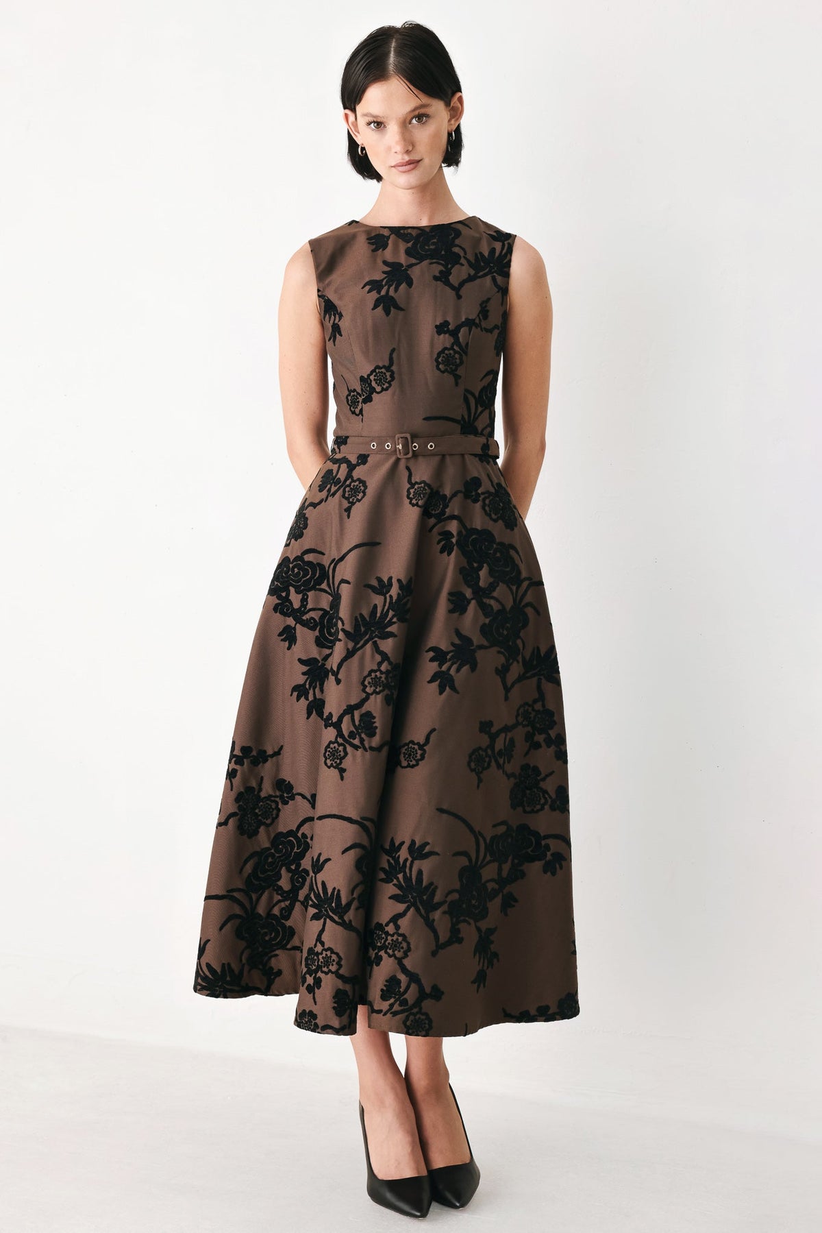 TRUFFLE BROCADE RICH BROWN MIDI DRESS | Lady Black Tie | A woman modeling a sleeveless brown midi dress with a black floral pattern and a matching belt, paired with black pointed-toe heels, against a plain white studio background.