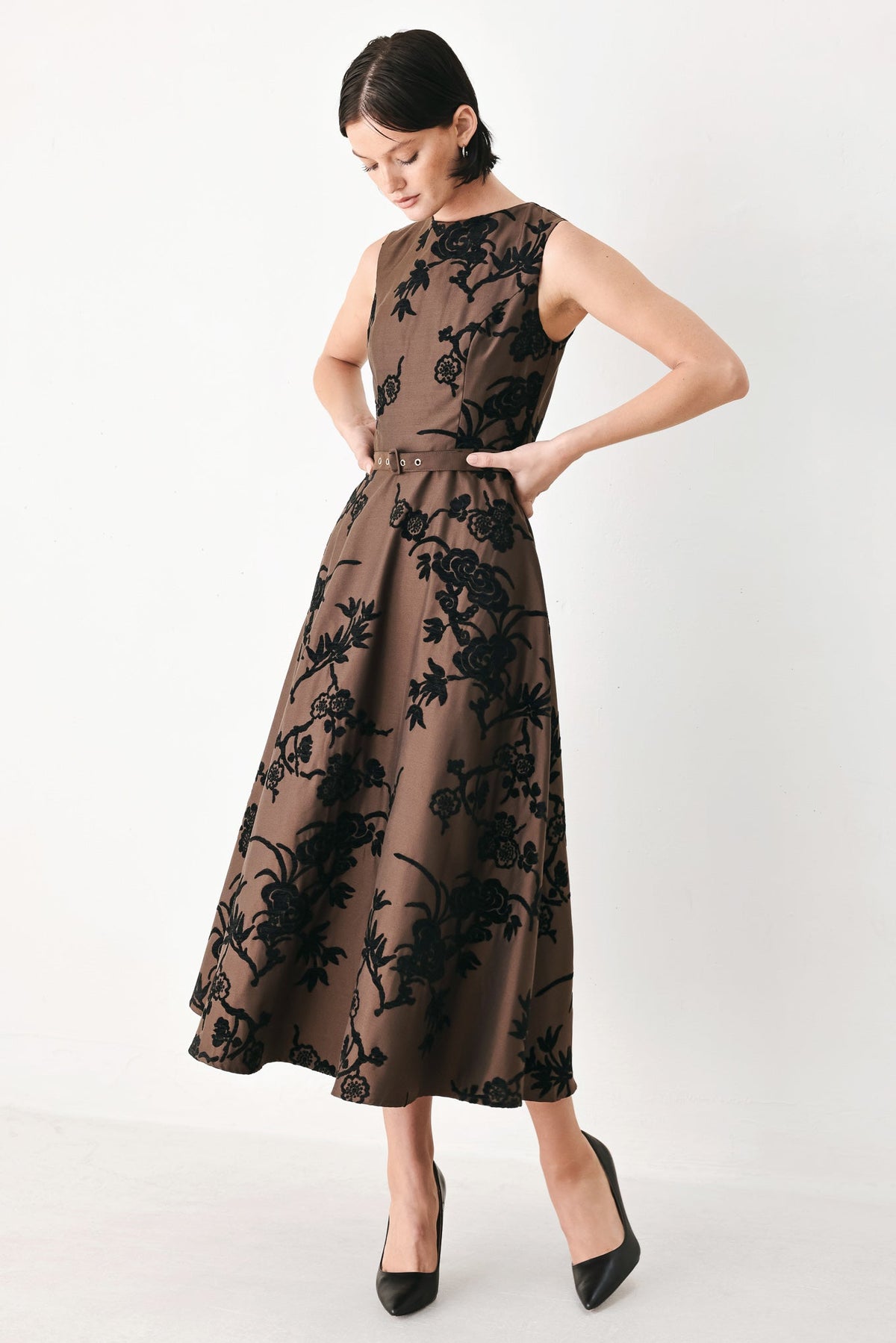 TRUFFLE BROCADE RICH BROWN MIDI DRESS | Lady Black Tie | A woman modeling a sleeveless midi dress with a brown base and black floral jacquard pattern, paired with black pointed-toe heels against a plain white studio background.
