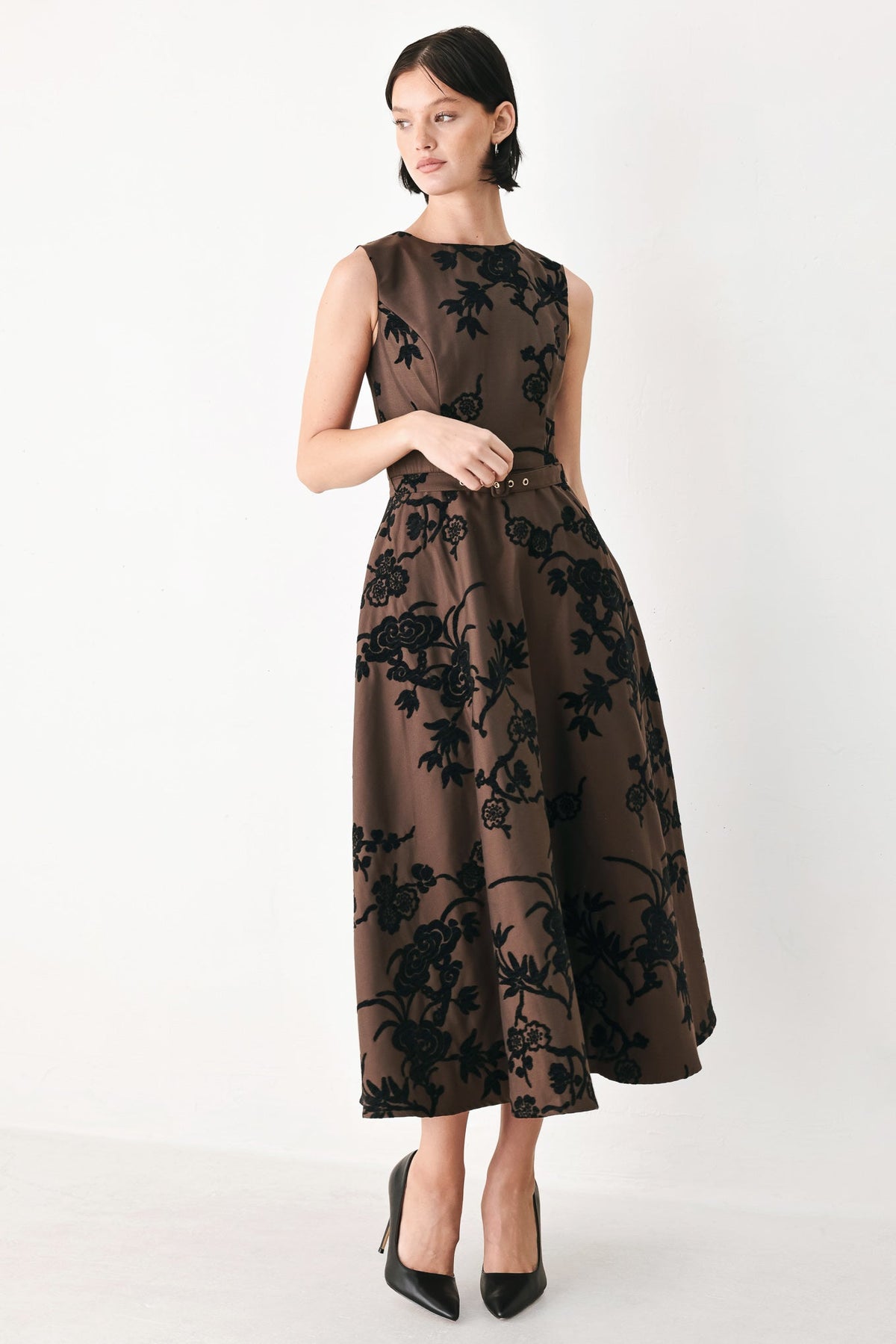 TRUFFLE BROCADE RICH BROWN MIDI DRESS | Lady Black Tie | A woman models a sleeveless midi dress with a brown base and black floral embroidery, paired with black pointed-toe heels against a plain white studio background.