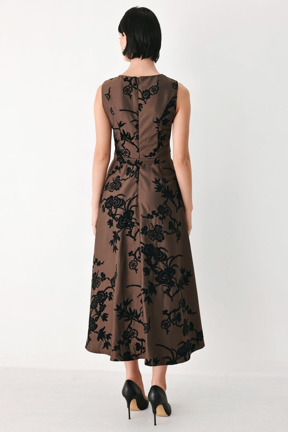 TRUFFLE BROCADE RICH BROWN MIDI DRESS | Lady Black Tie | A woman viewed from the back wearing a sleeveless brown midi dress with a black floral jacquard pattern and black heels, set against a plain white studio background.
