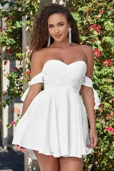 A woman wears a white, strapless dress with bow-tied sleeves, standing confidently among lush greenery and flowers in a bright outdoor setting, exuding a cheerful and elegant vibe.